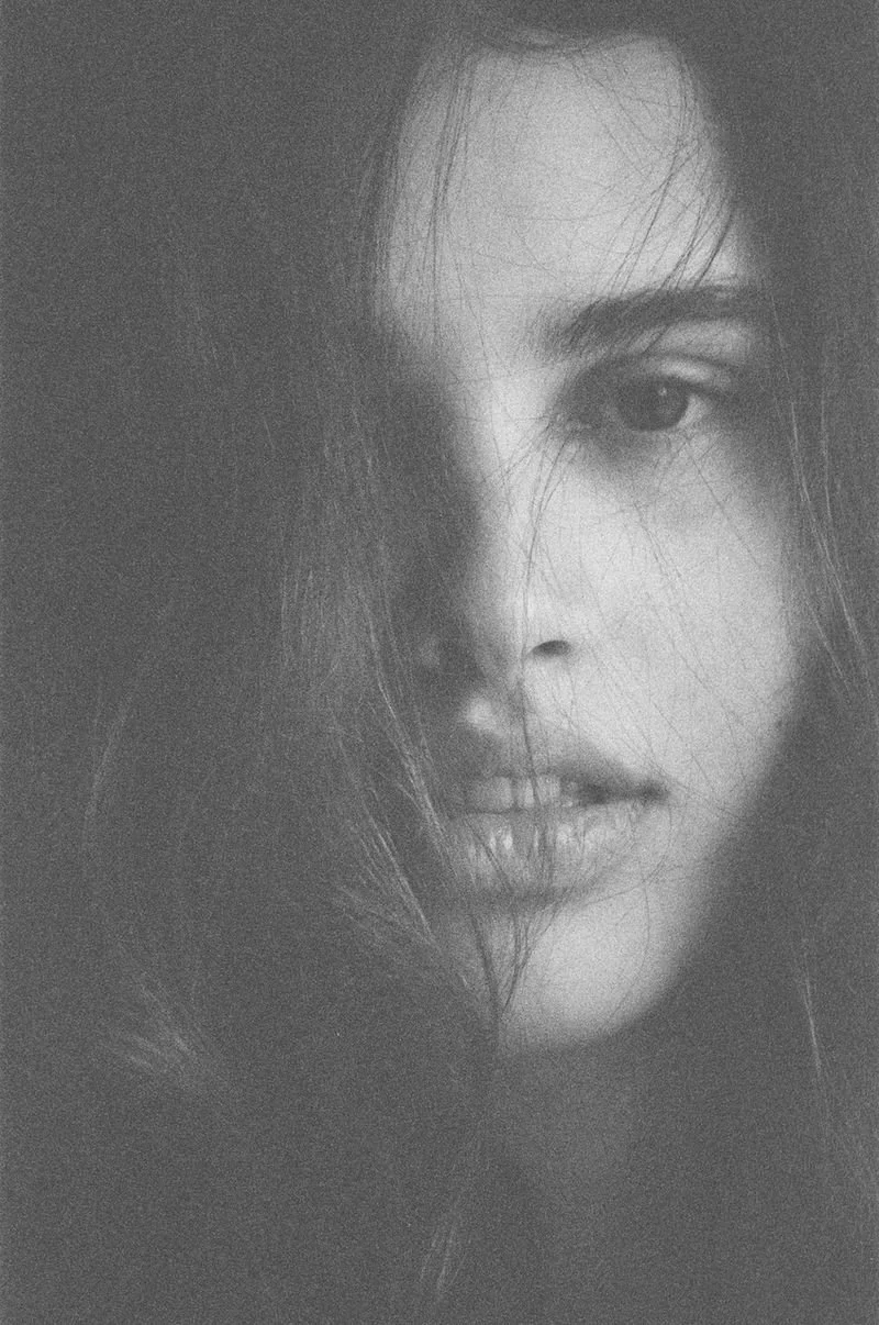 Close-up black and white portrait of a woman's face, with some hair partially covering her face, one eye visible, showing a neutral expression.