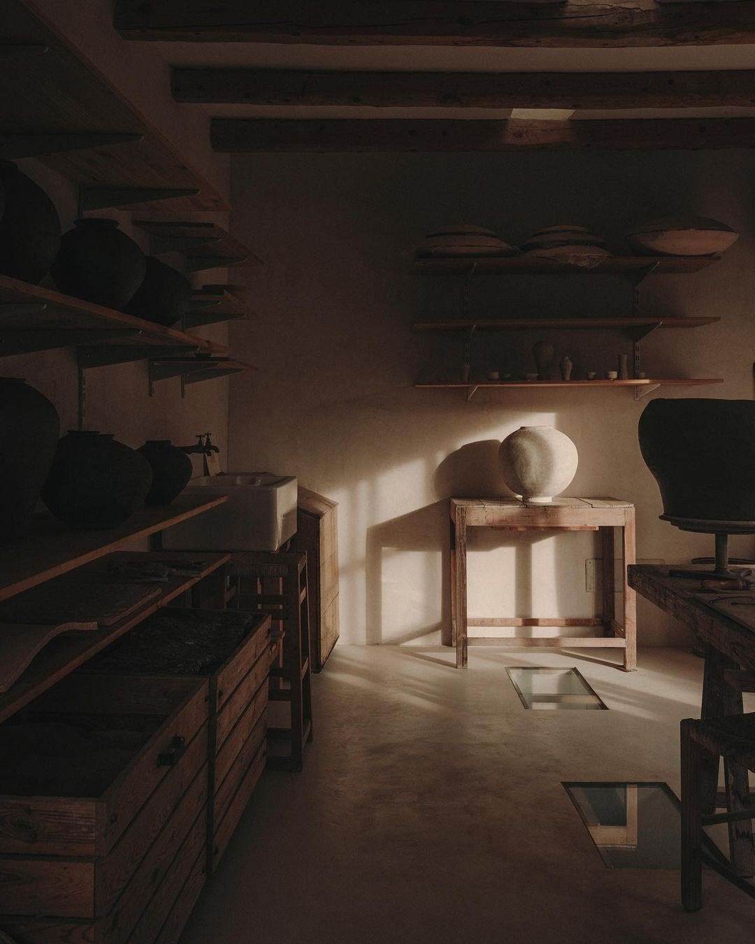 A dimly lit room with shelves of pottery, a wooden table with a large pottery piece, and a black office chair.