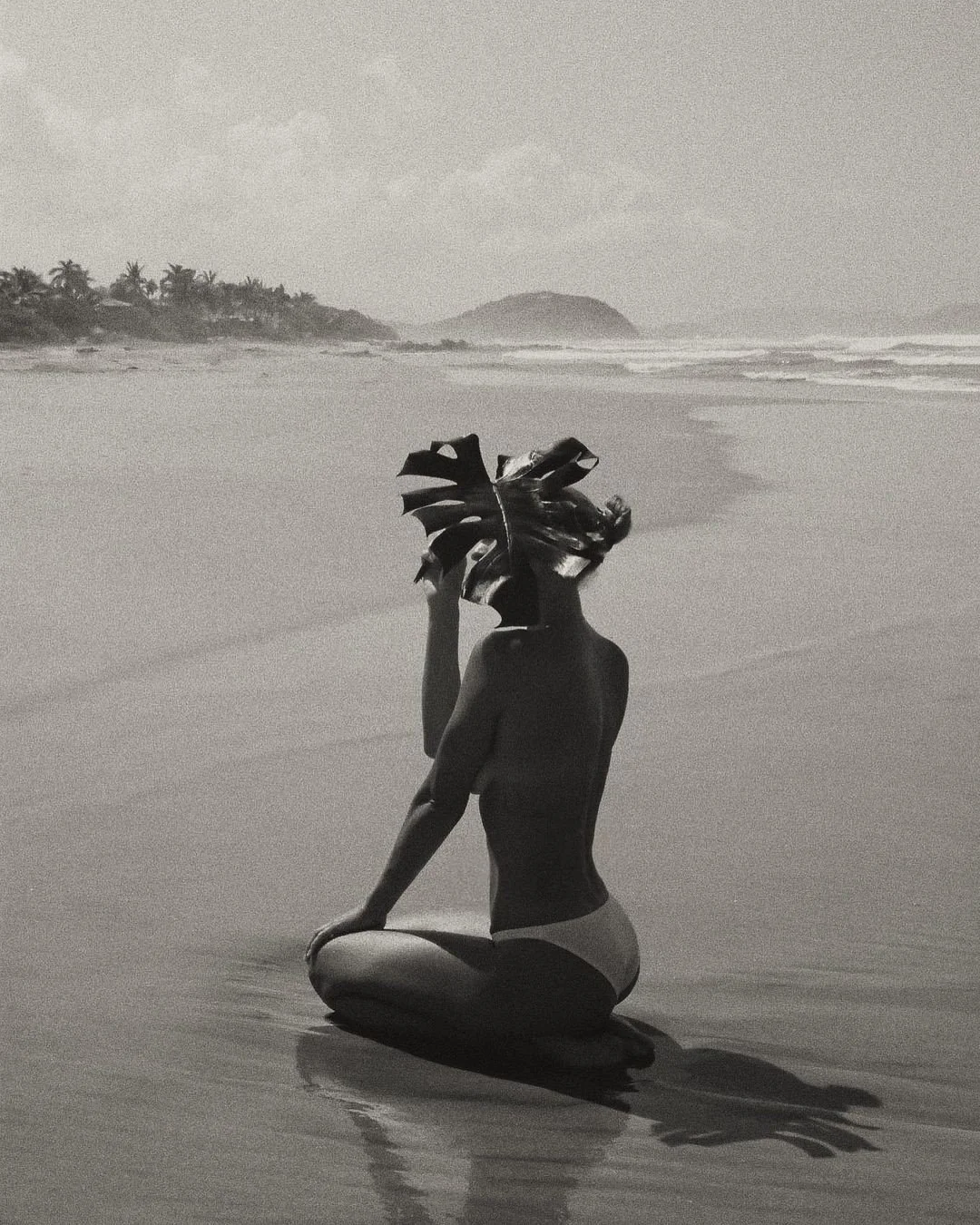 A person sitting cross-legged on the beach, holding a large leaf in front of their head, with an ocean, surf, and distant land in the background in black and white.