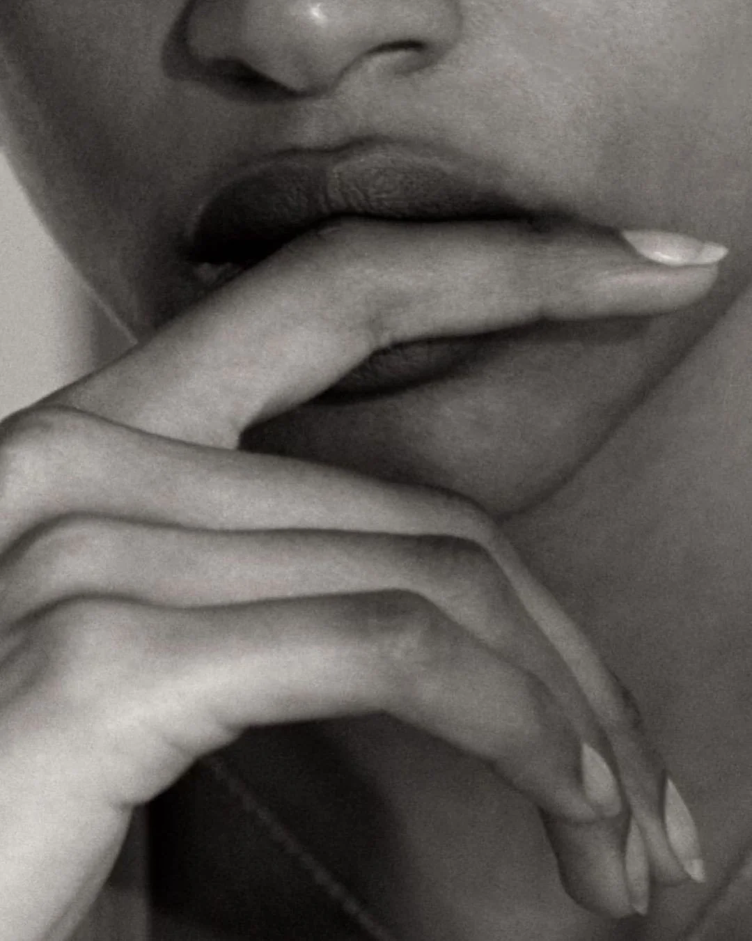 Close-up black and white photo of a person's lips, nose, and hand touching lips with the index finger, with the thumb on the chin.