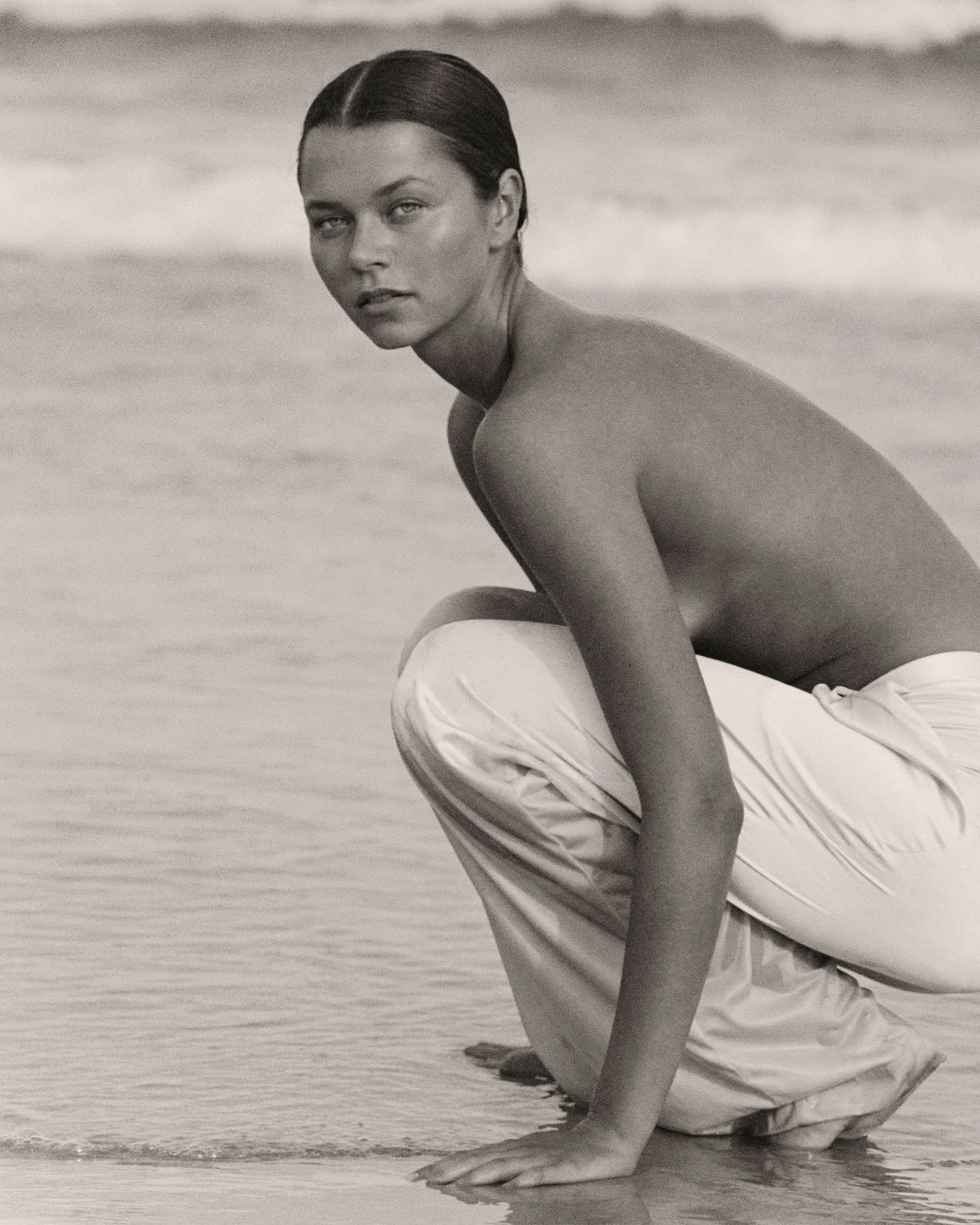 A woman crouches on the beach in front of the water, wearing white pants, with wet hair, looking at the camera.