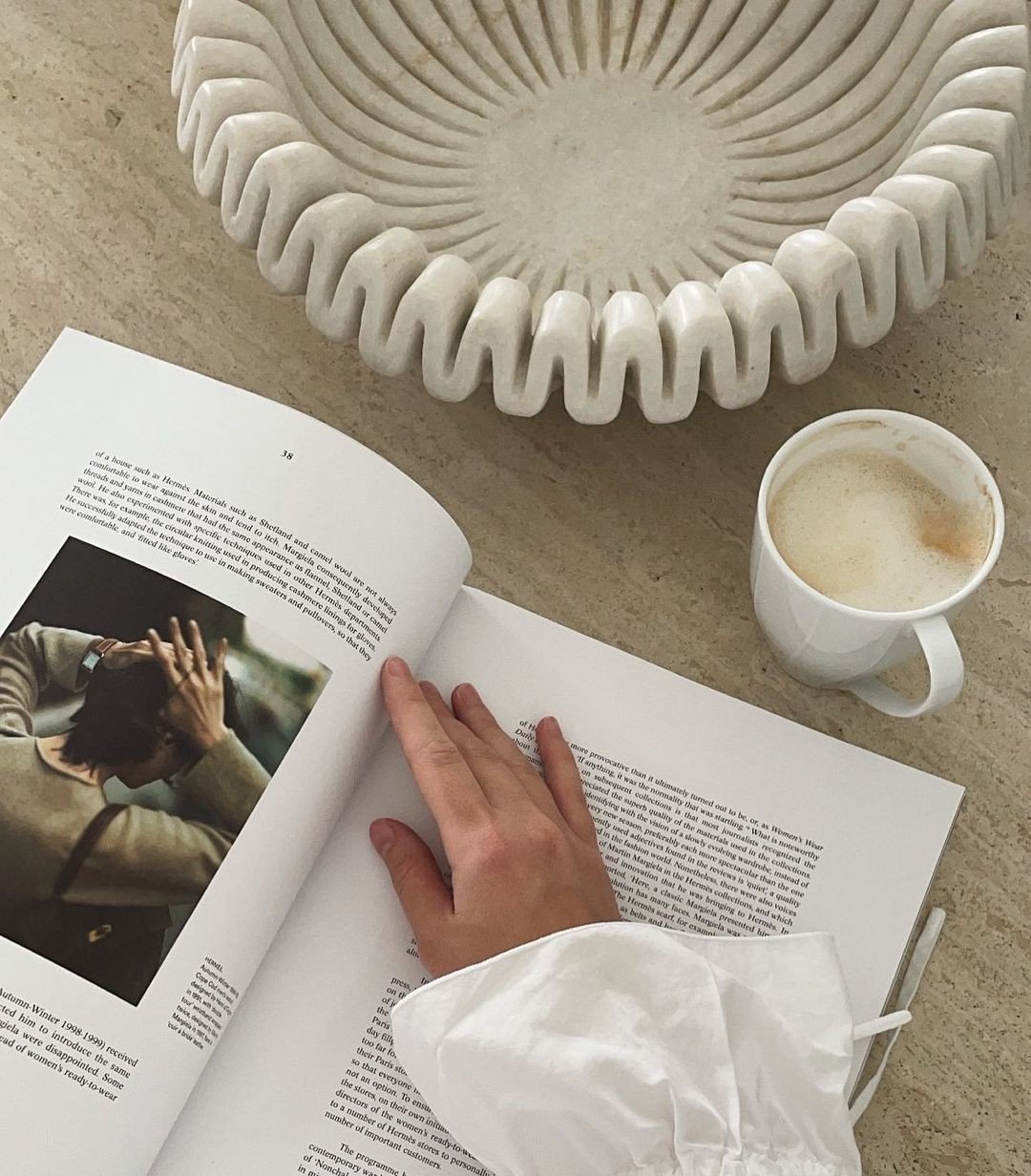 A person reading a magazine at a table with a white coffee mug and a decorative white bowl with ridges, on a beige tabletop.