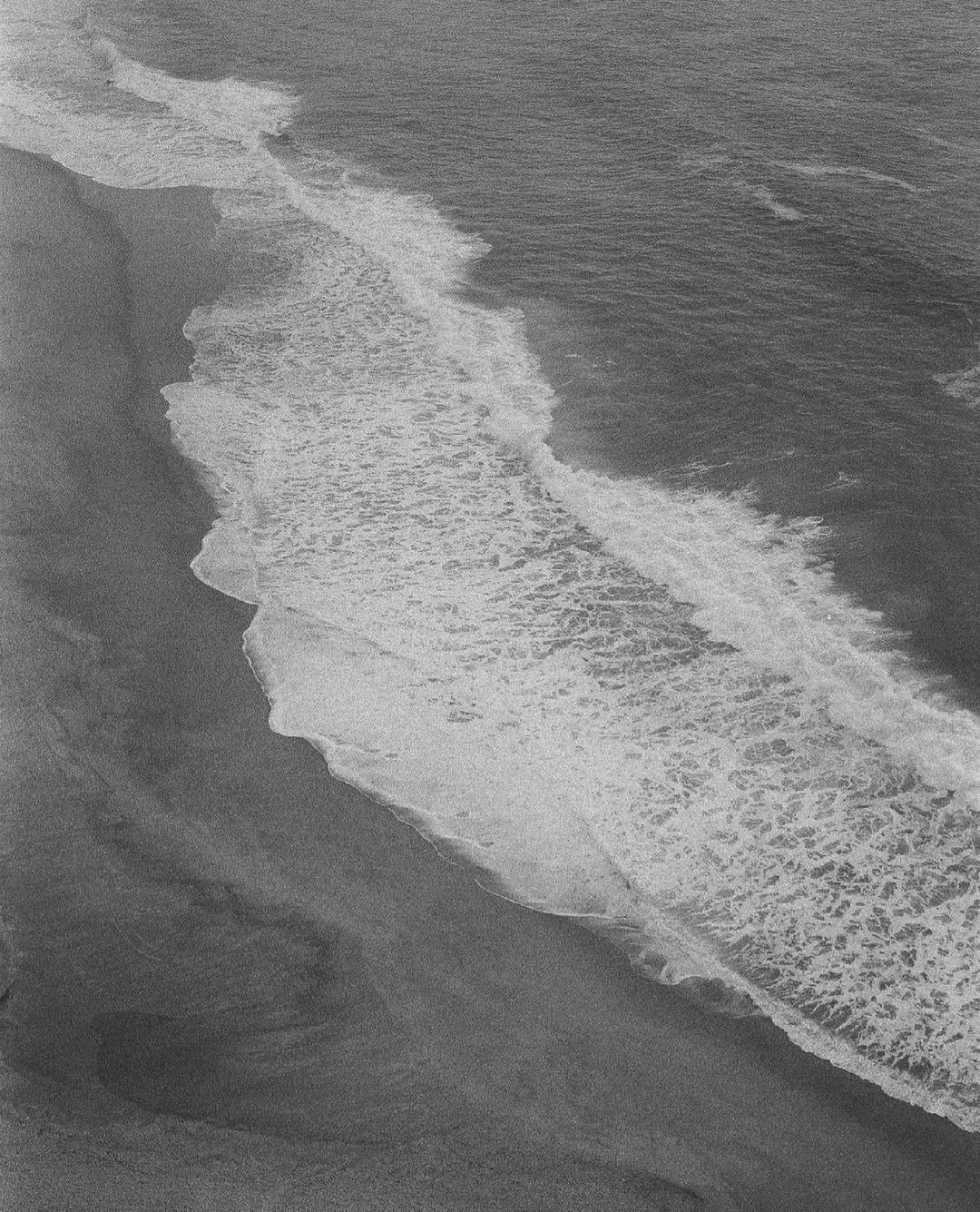 Black and white aerial view of the ocean shore with waves crashing onto the sandy beach.