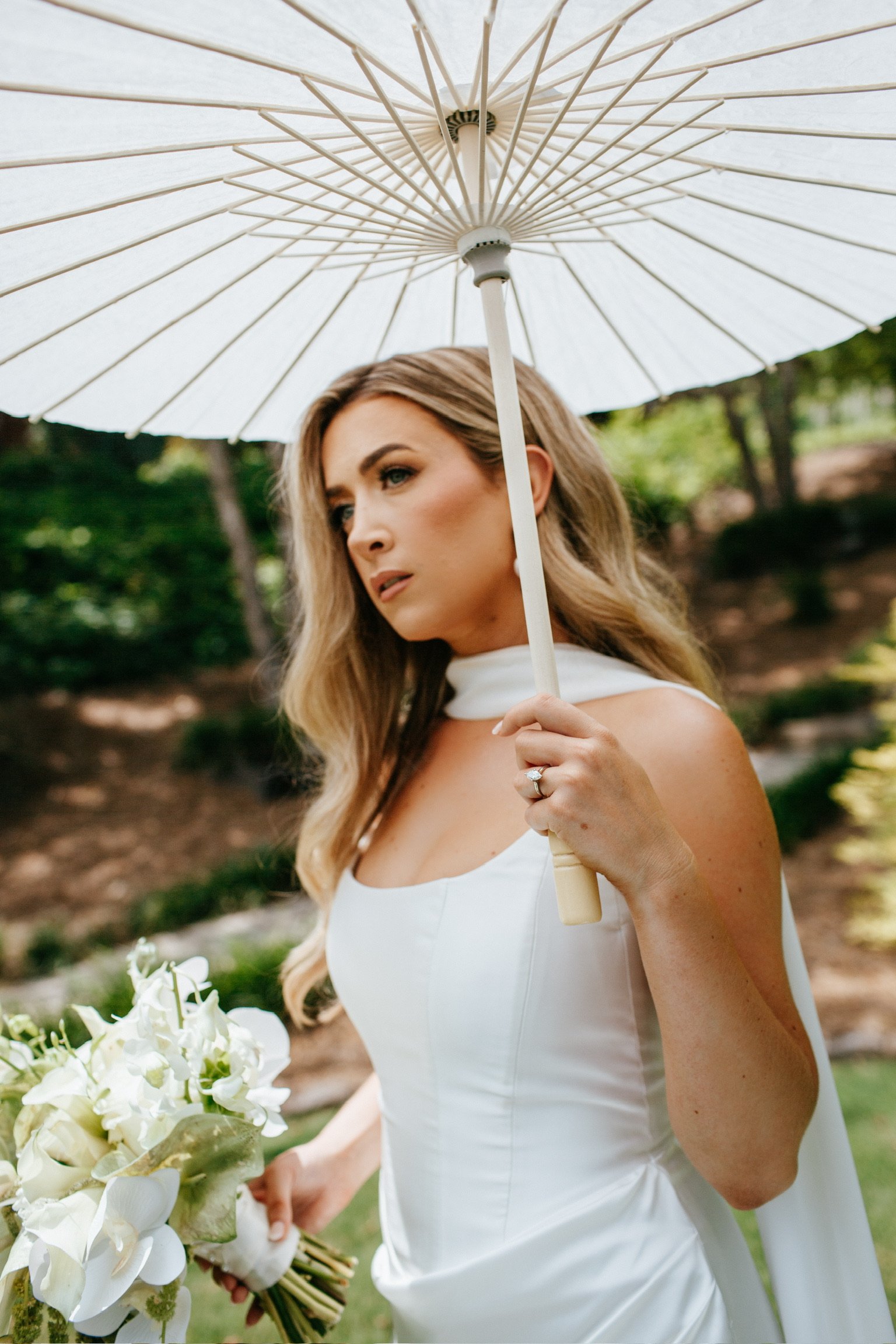 A woman in a white wedding dress holds a bouquet of white flowers in one hand and an open white umbrella in the other, standing outdoors in a garden setting.