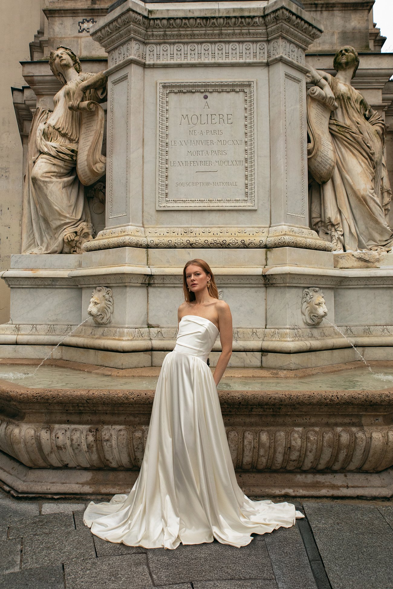 A woman in a long white strapless gown stands in front of a marble fountain with intricate carvings and statues, in front of a large stone monument with inscriptions.