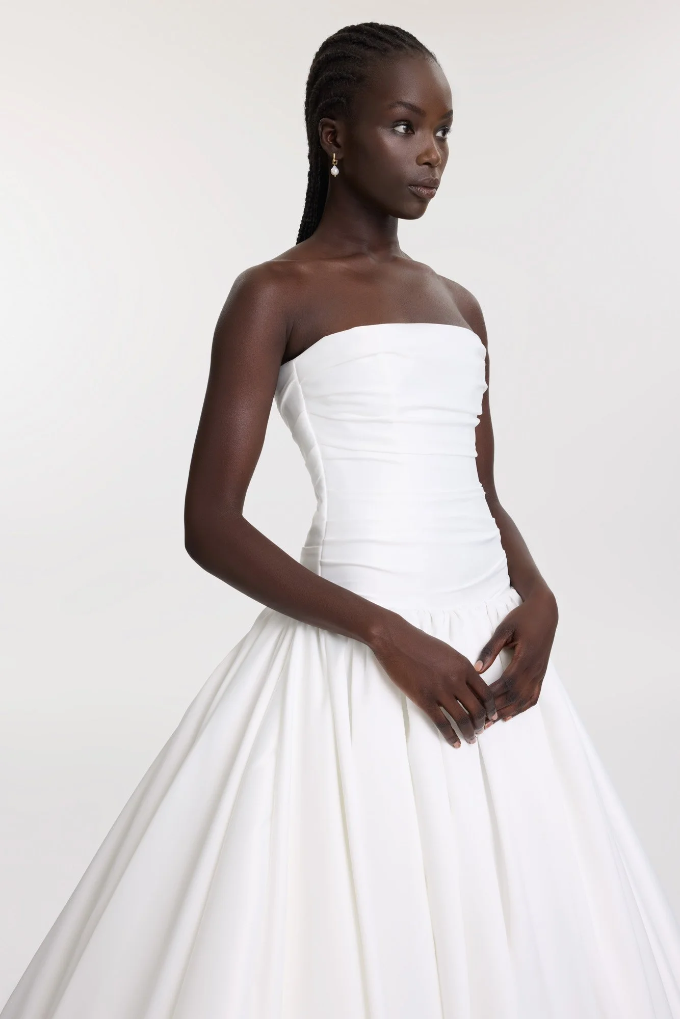 A woman with dark skin and braided hair wearing a white strapless wedding gown with a fitted bodice and a full skirt, standing against a plain white background.