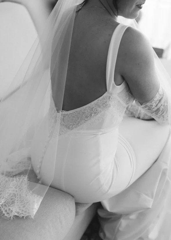 Woman in a wedding dress sitting on a couch, with her back partially visible and her veil flowing over her shoulder.