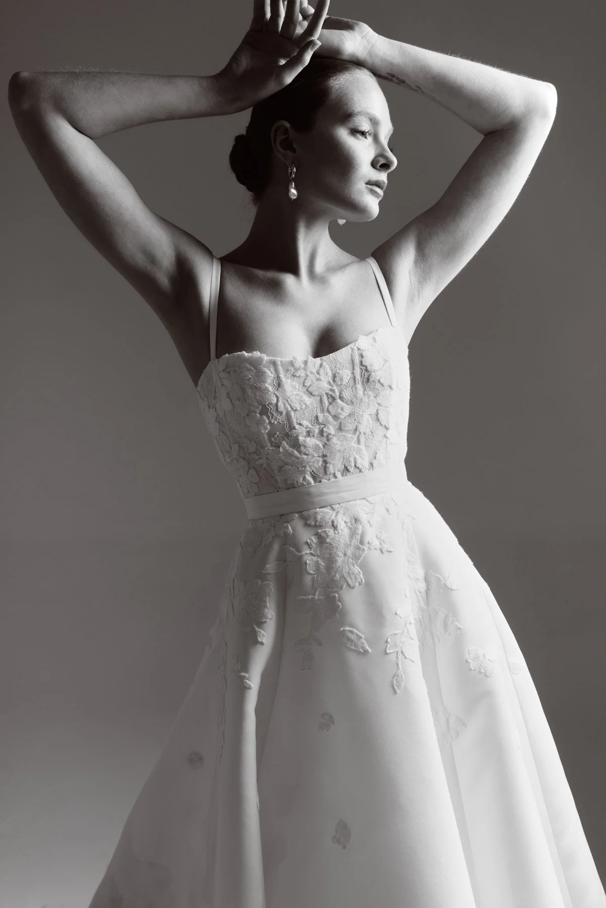 A woman wearing a white lace wedding dress, posing with her arms raised above her head, in profile view in a black and white photo.