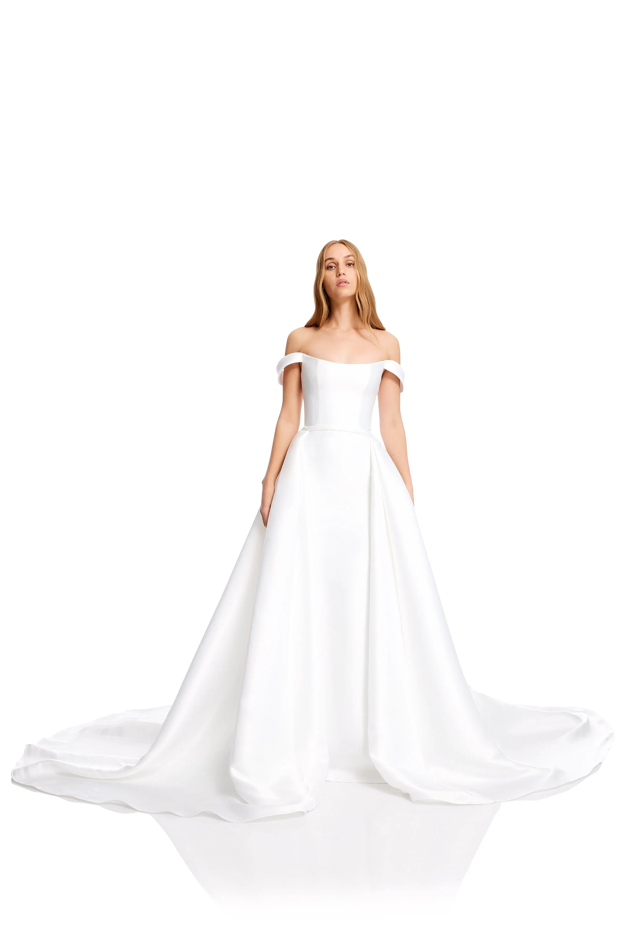 A woman wearing an elegant off-the-shoulder white wedding gown with a full skirt standing against a white background.