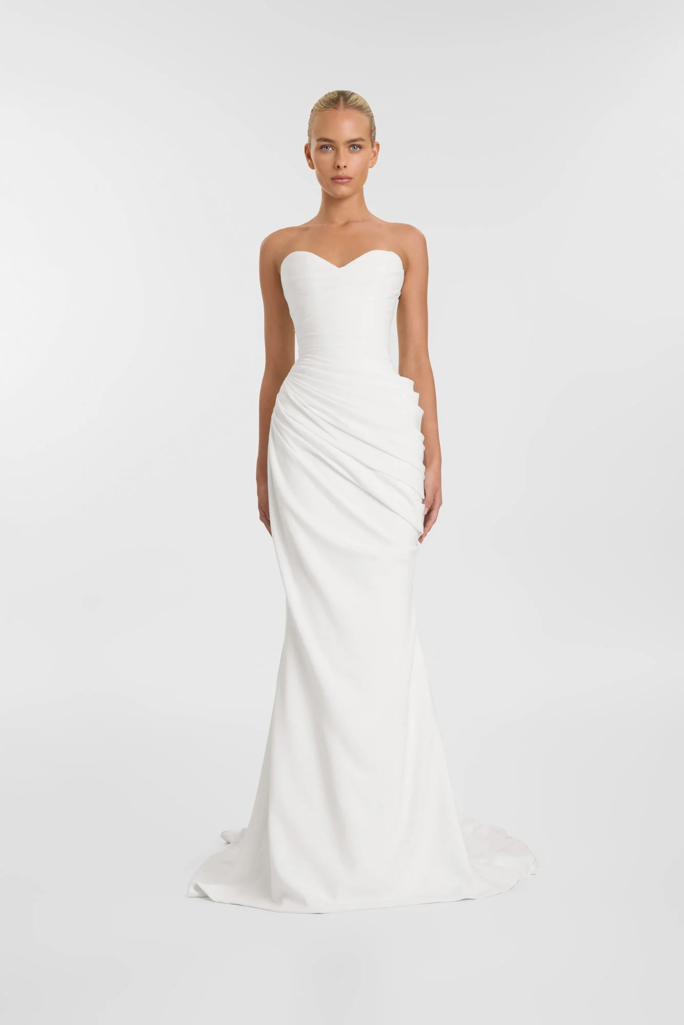 A woman wearing a strapless white wedding dress with a fitted bodice and a long, gathered skirt standing against a plain white background.