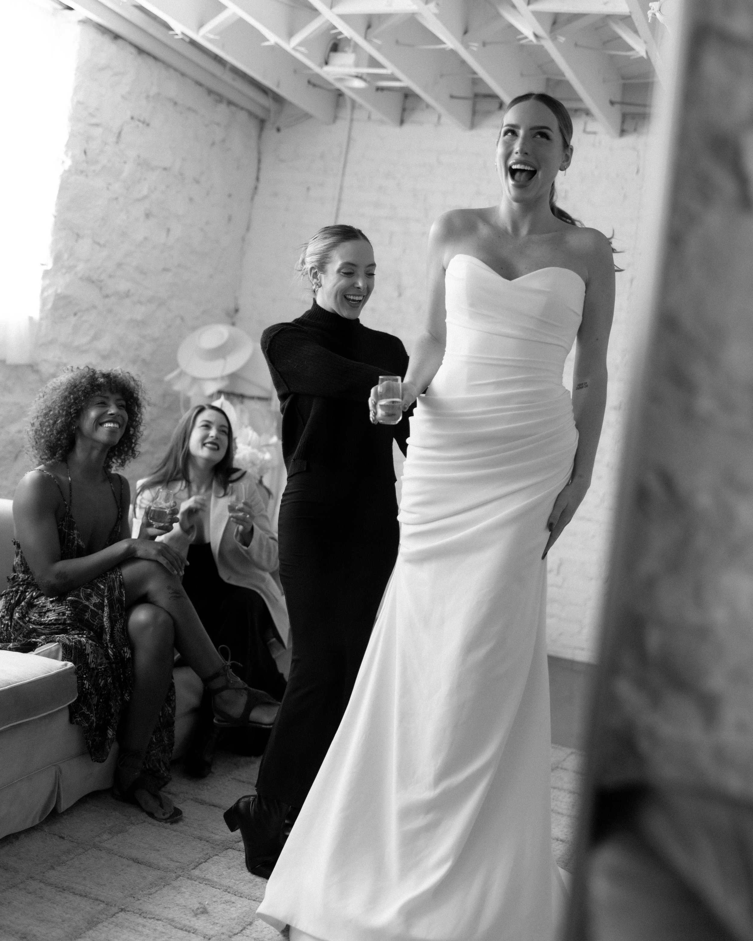 A bride in a strapless wedding dress laughing with women at a celebration, with some women holding drinks, in a cozy room with brick walls and a high ceiling.