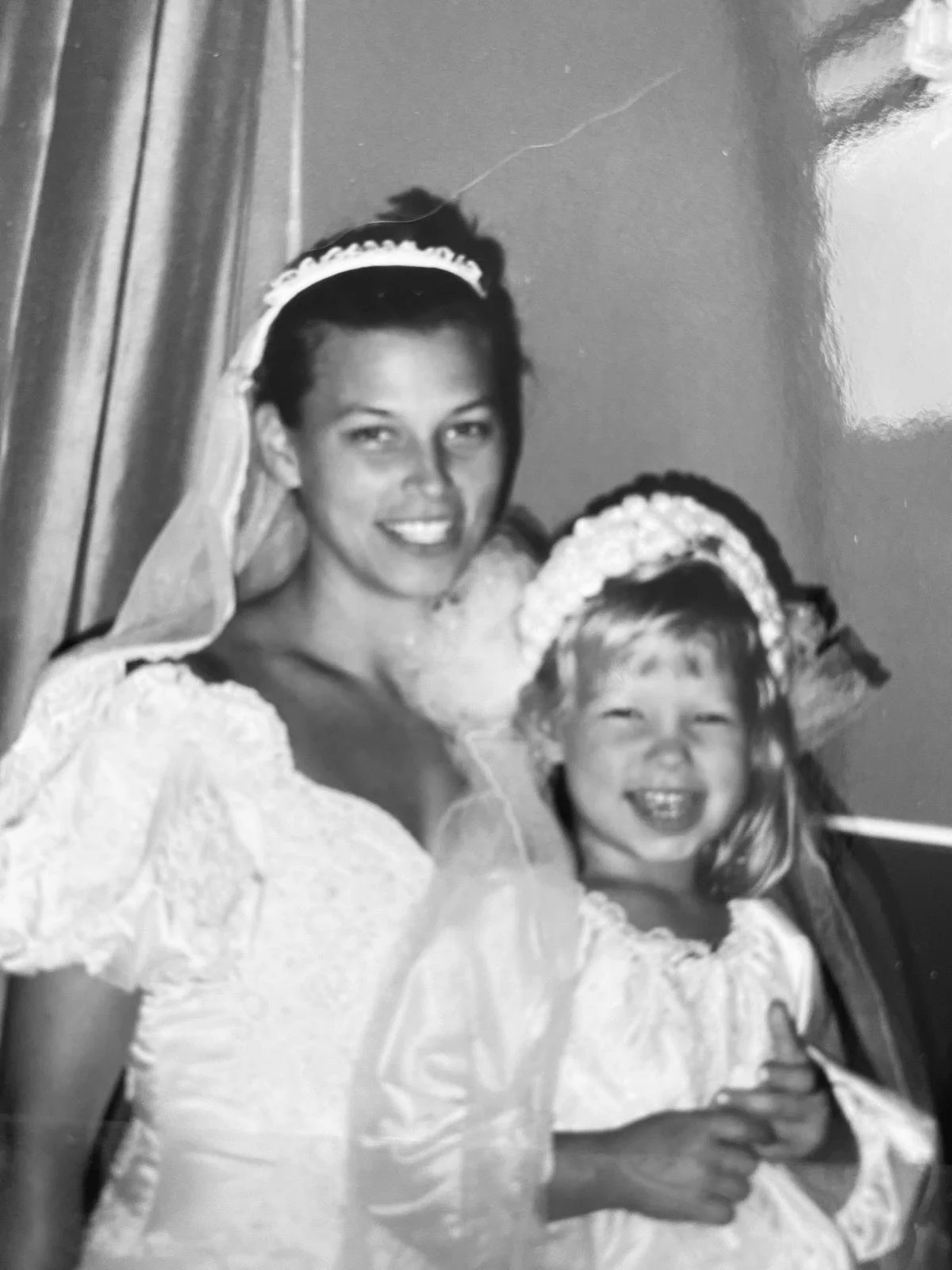 A woman in a wedding dress with lace sleeves, wearing a veil, stands next to a young girl in a dress with puffed sleeves, a floral headband, and a big smile.