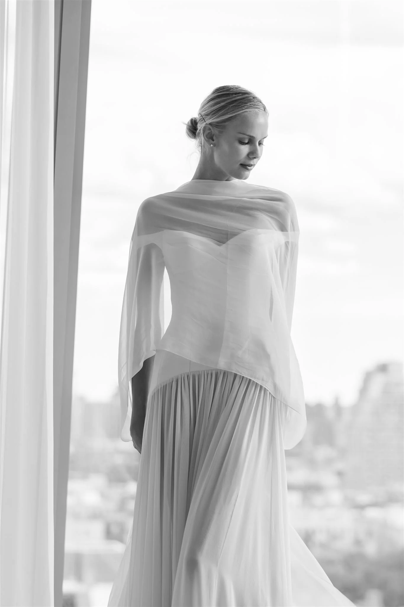 Black and white photo of a woman in a flowing dress standing by a window, looking down.