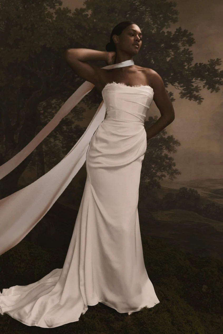 A woman in a strapless white gown with a singe shoulder strap pose with one hand behind her head against a painted landscape background.