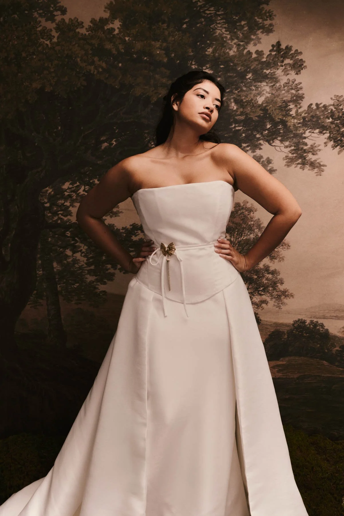A woman in a strapless white dress with a butterfly brooch stands with her hands on her hips in front of a mural of trees and landscape.