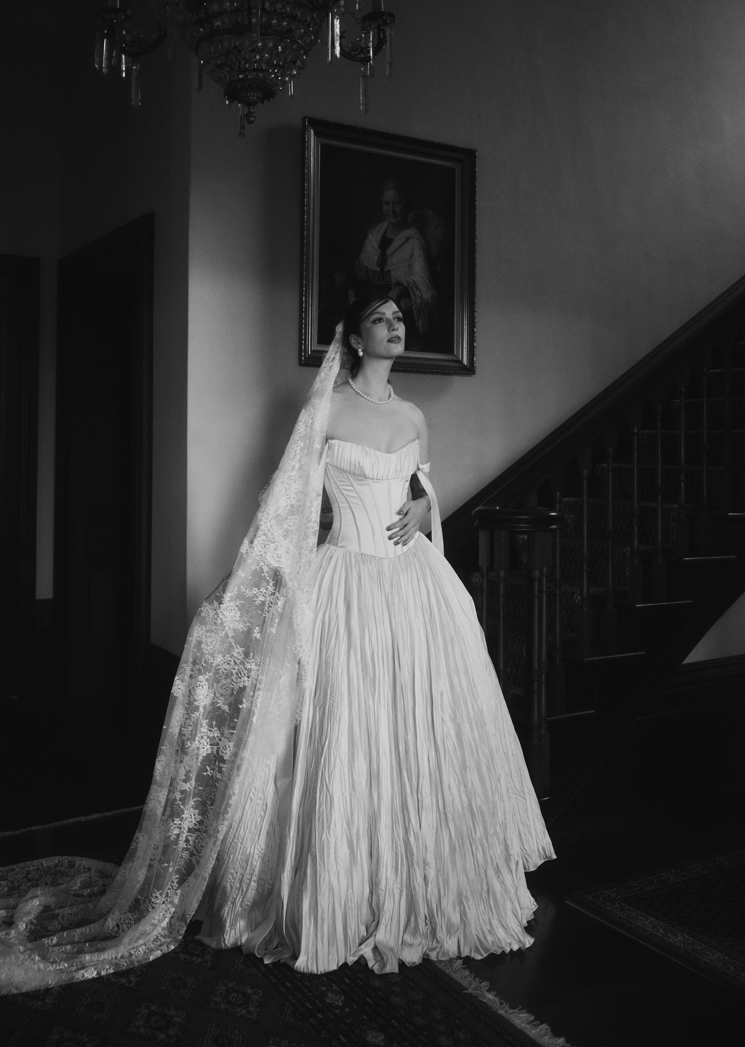 A woman in an elegant wedding dress standing indoors next to a staircase, with a portrait hanging above her, in black and white.