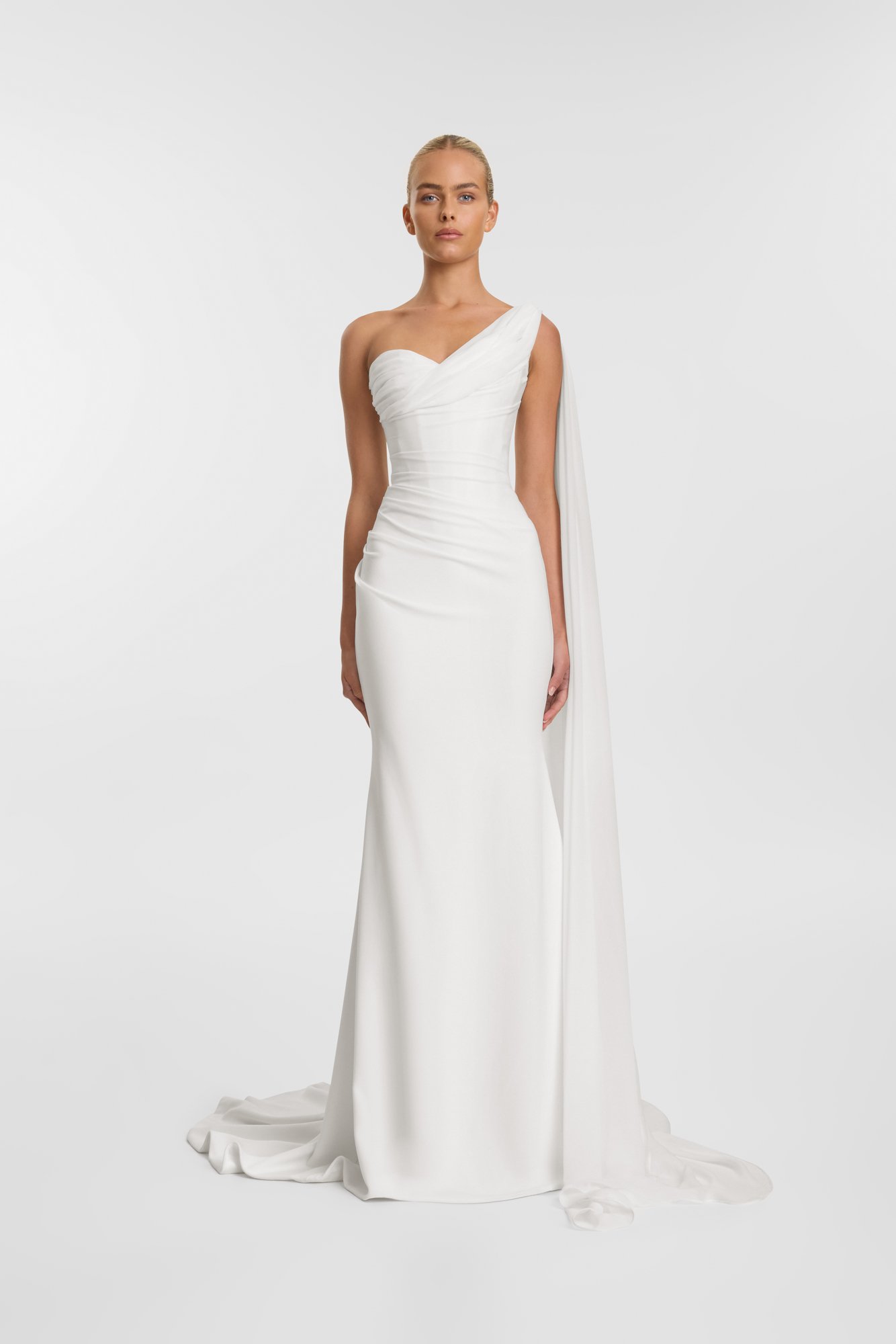 A woman modeling a white, asymmetrical wedding gown with one shoulder and a long train.