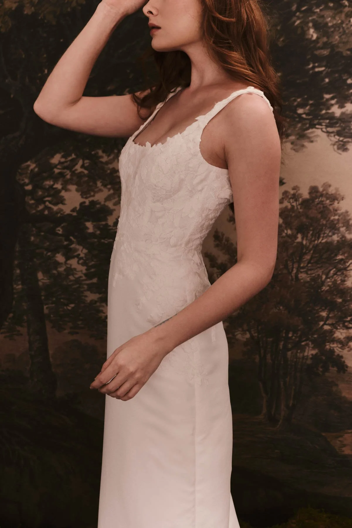 Side view of a woman wearing a white lace sleeveless dress with a tree background.