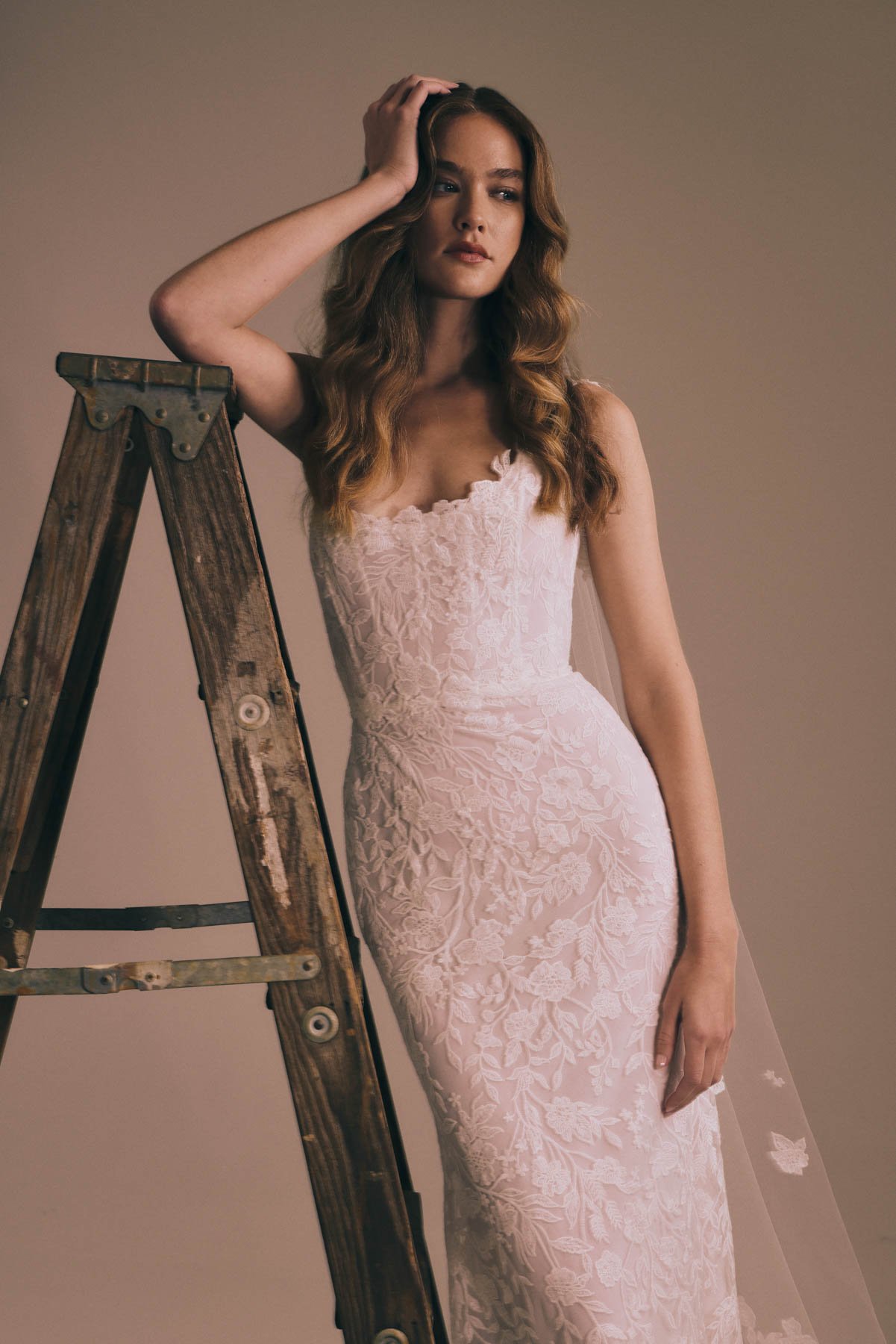 A woman in a pink lace wedding dress standing next to a wooden ladder, touching her hair with her right hand, looking thoughtful.