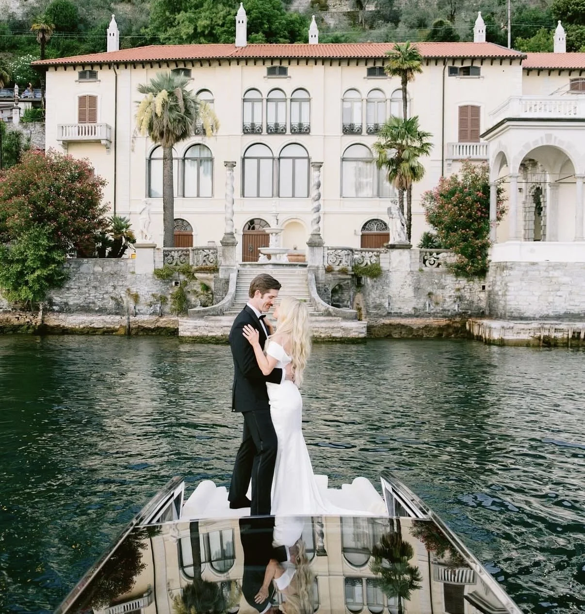 A couple in wedding attire dancing on a boat on a waterway, with a grand mansion and trees in the background.