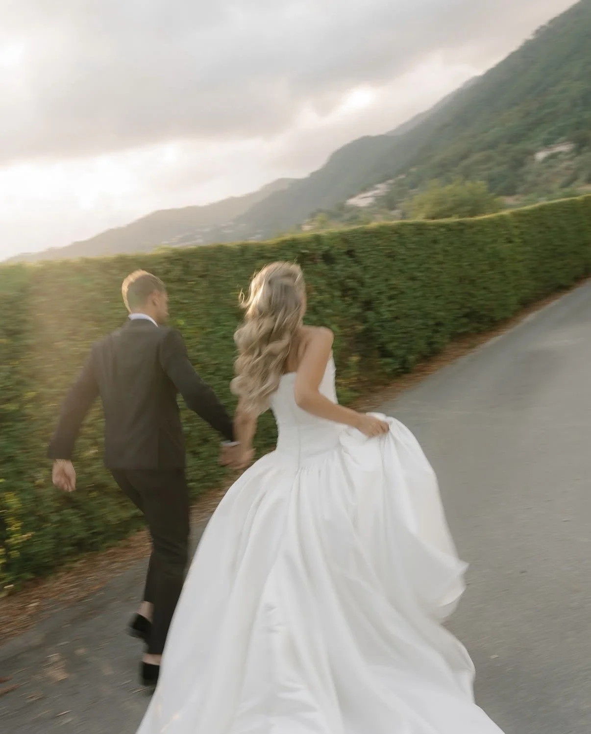 A bride and groom holding hands and running on a road surrounded by green hedges and mountains, with cloudy skies overhead.
