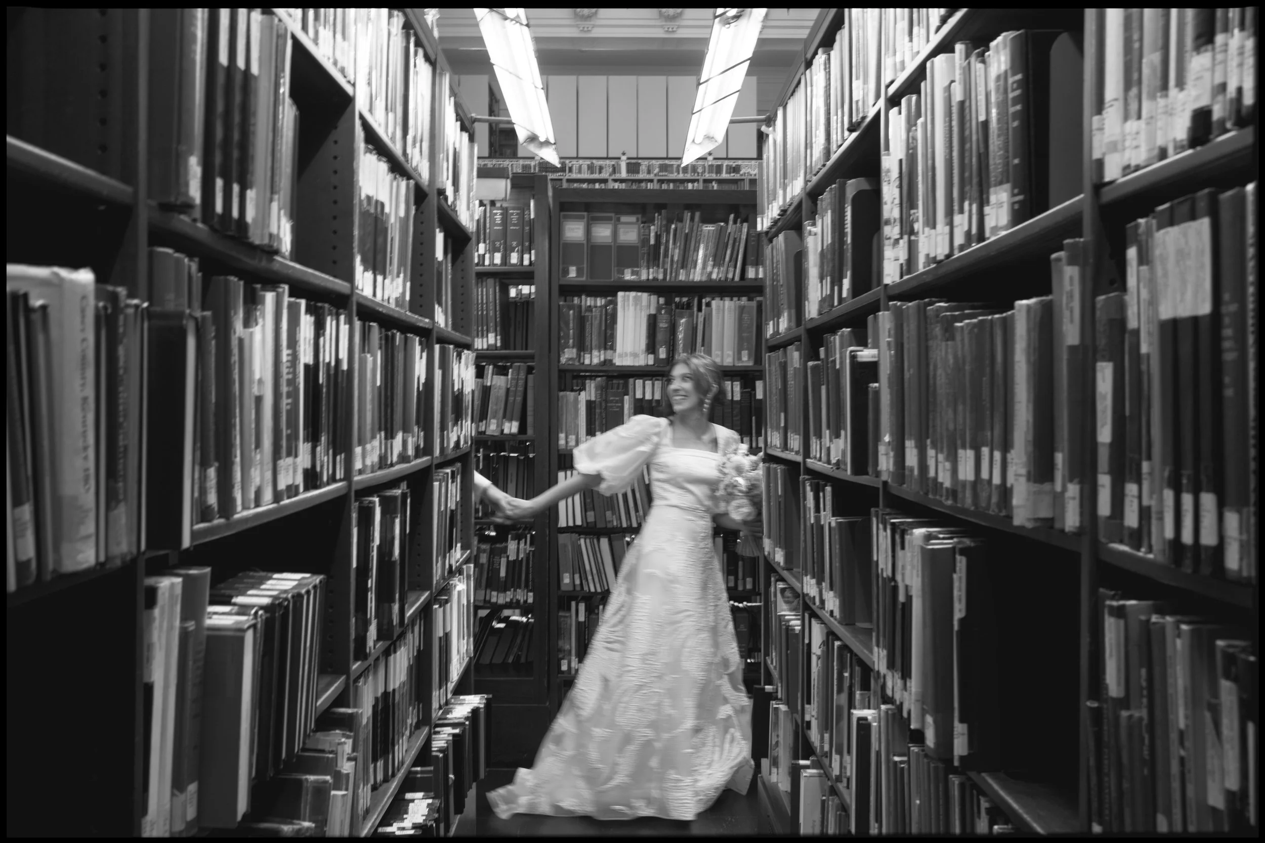 A woman in a long, elegant dress holding a bouquet of flowers is browsing books in a library aisle filled with tall shelves of books. The photo is in black and white.