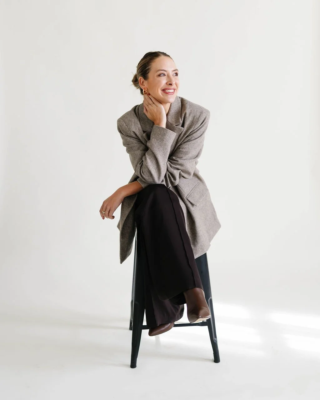 A woman sitting on a black stool, smiling with her hand on her neck, wearing a gray blazer, black pants, and brown shoes, against a plain white background.