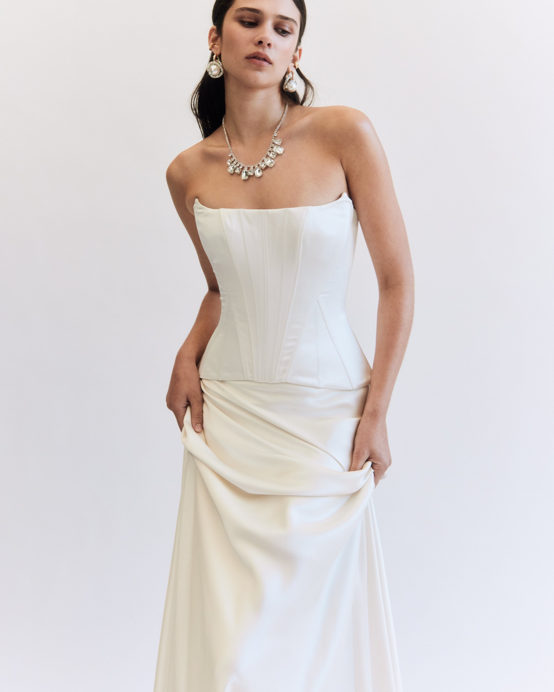 A woman wearing a strapless white wedding gown with pearl jewelry and holding part of her dress.