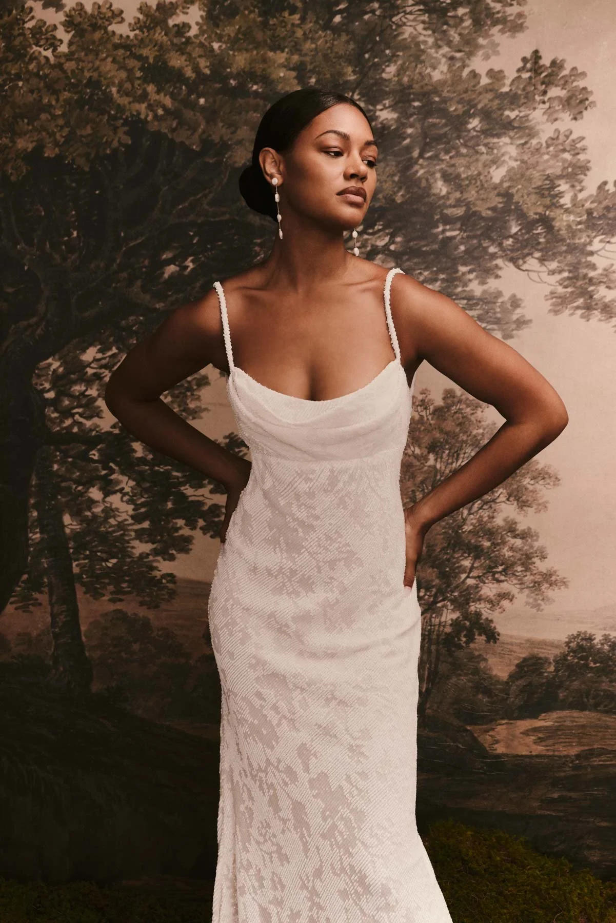 A woman in a white, elegant dress with spaghetti straps stands in front of a painted backdrop of trees and landscape, posing with hands on her hips and looking to the side.