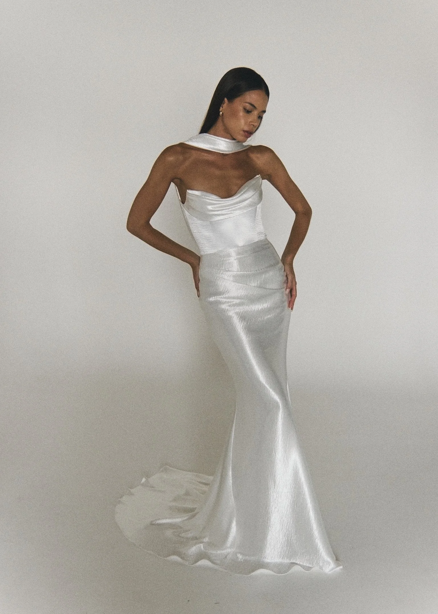 Woman in a shiny white satin gown with an asymmetrical top and a mermaid skirt, standing against a plain white background, with her hands on her hips and looking down.