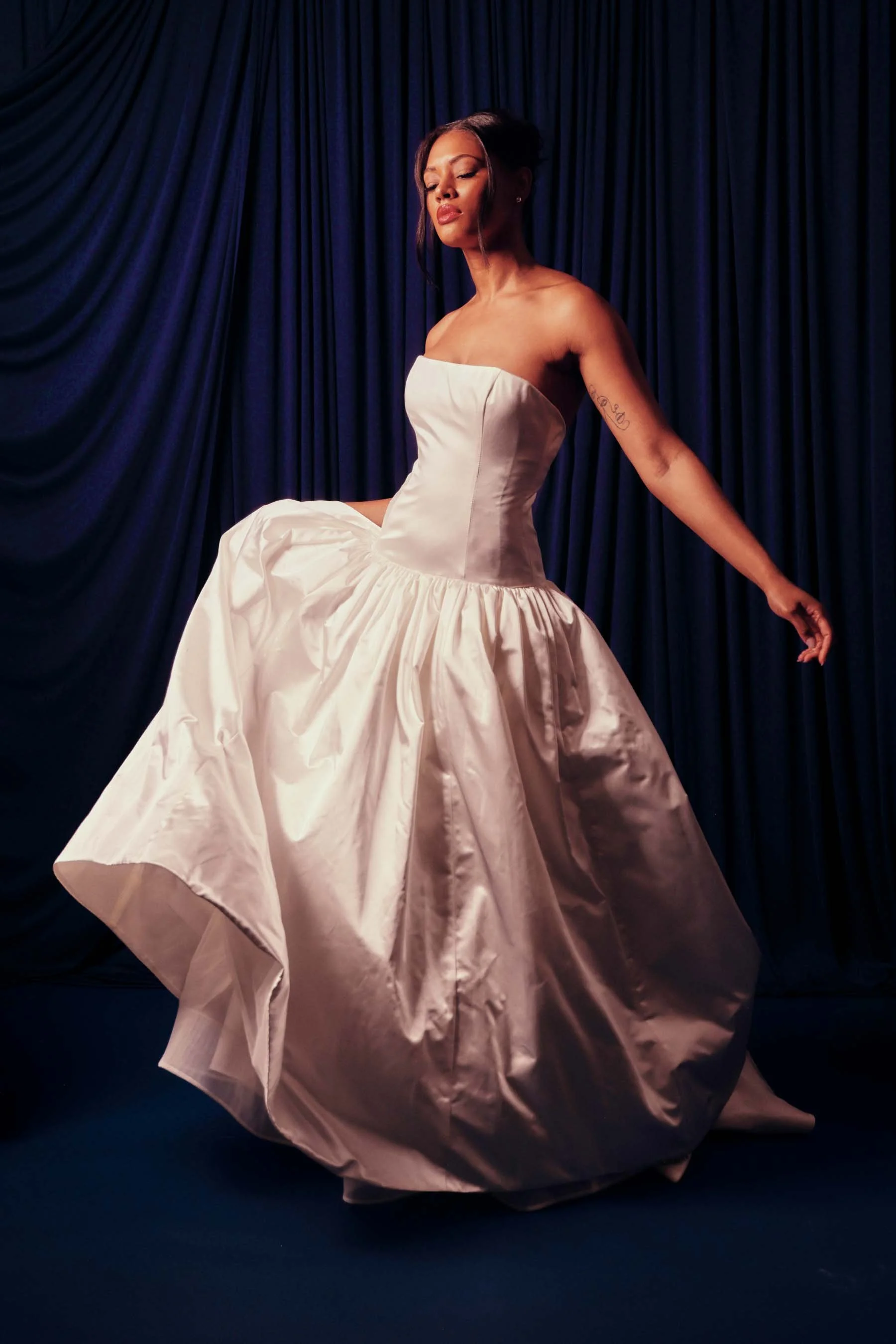Woman in a strapless white satin ball gown dancing in front of a dark blue curtain.