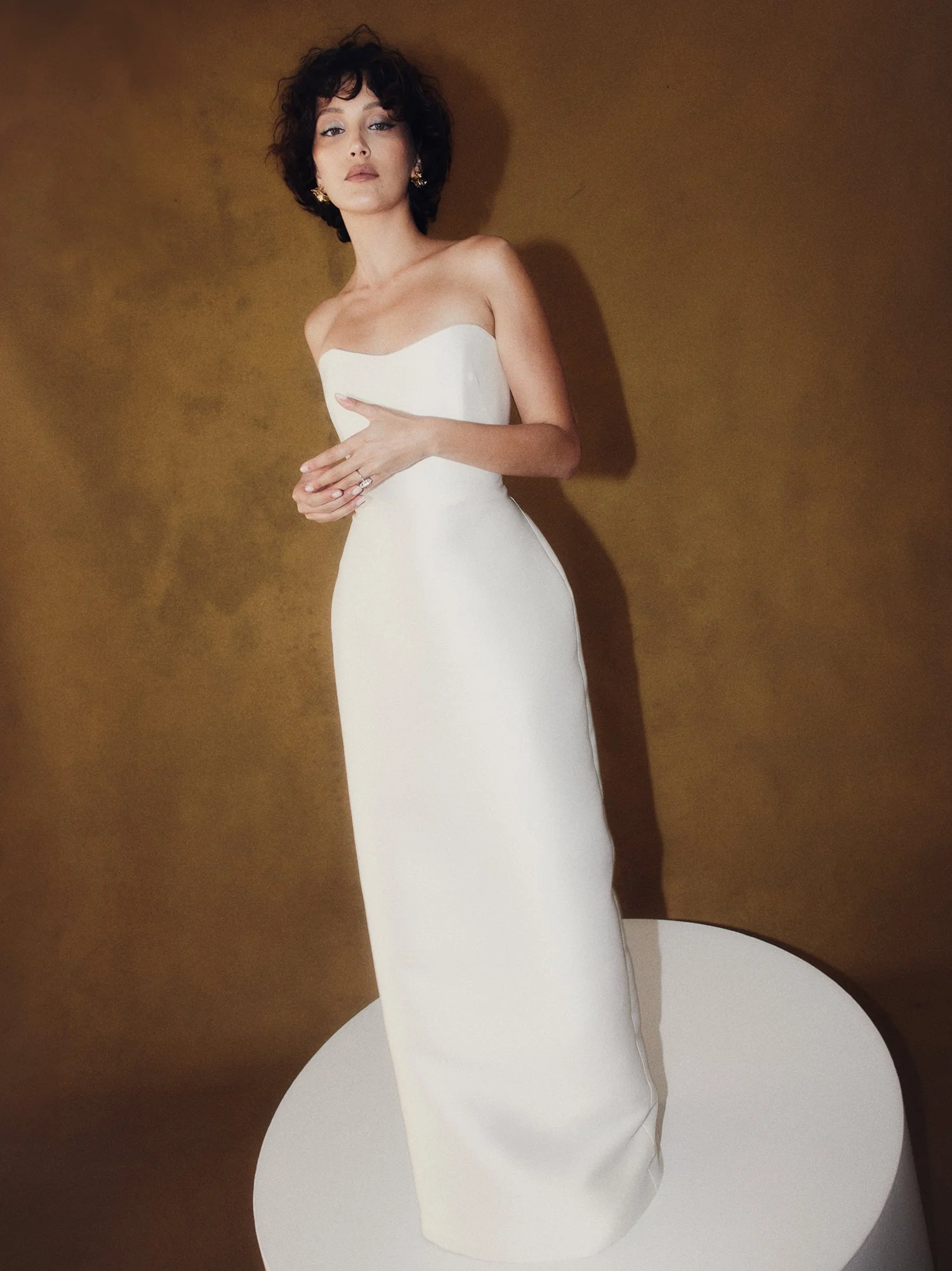 A woman with short curly dark hair and earrings wearing a strapless white gown, standing on a white circular platform against a brown background.