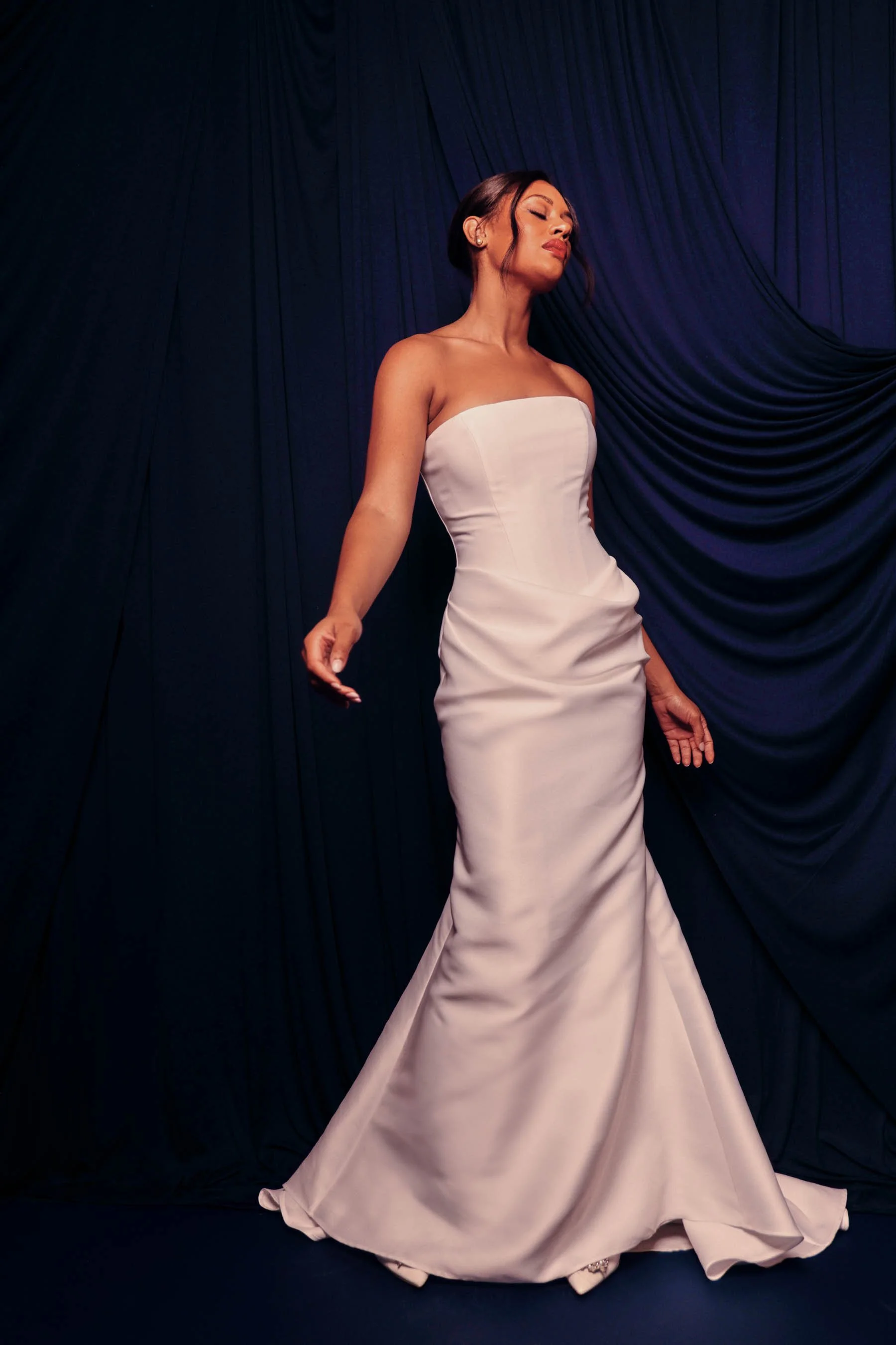A woman in a strapless white evening gown standing against a dark blue curtain, posing with her eyes closed and head tilted slightly to the side.