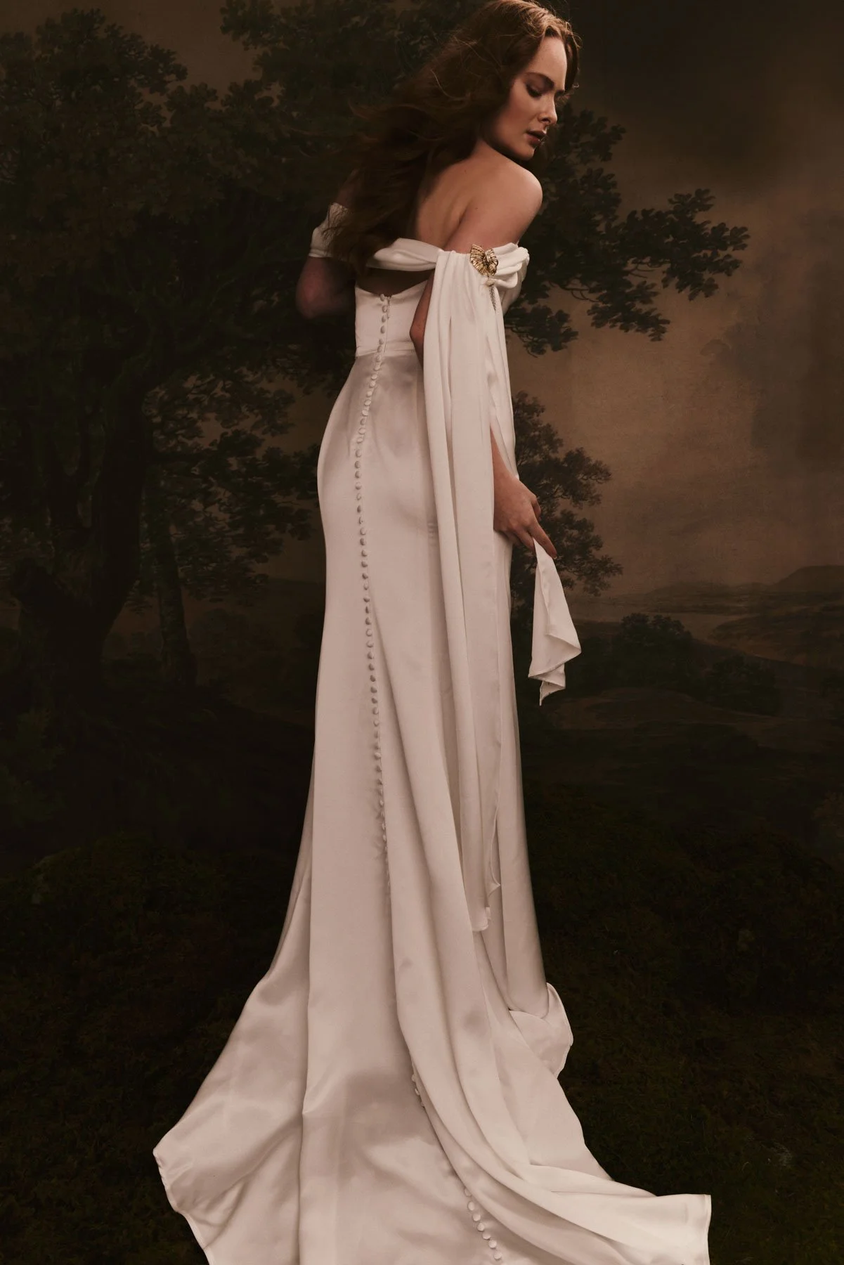 A woman in an elegant off-shoulder wedding dress with buttons down the back, standing outdoors with a dark, cloudy sky and trees in the background.