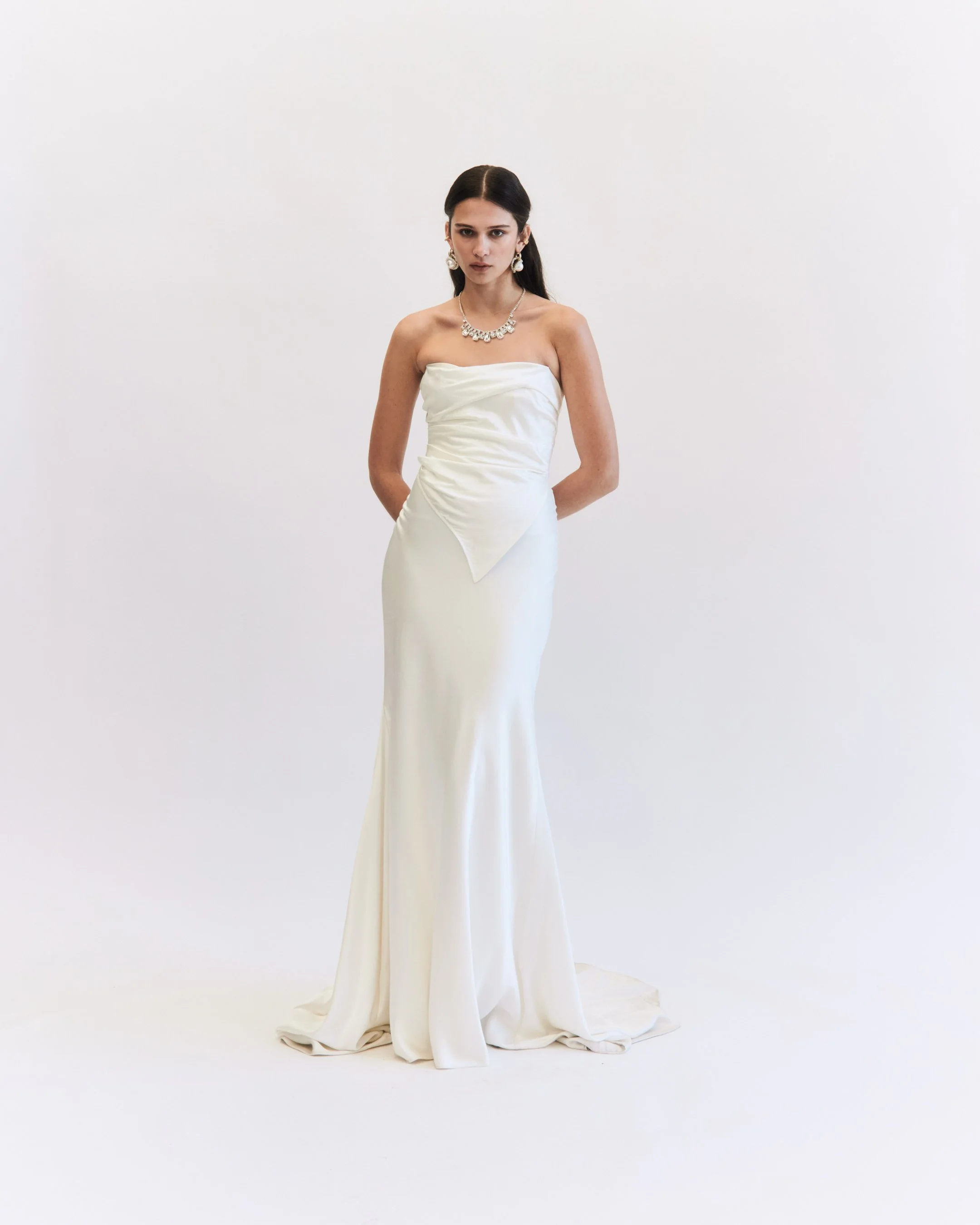 A woman in a strapless white wedding dress with jewelry, standing against a plain white background.