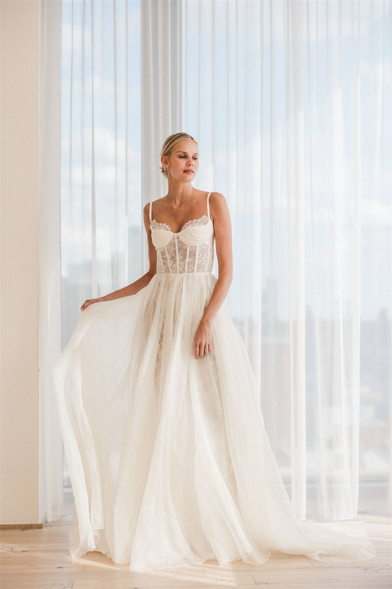 A woman in a white wedding dress standing in front of sheer curtains, holding the skirt with one hand, looking off to her right.