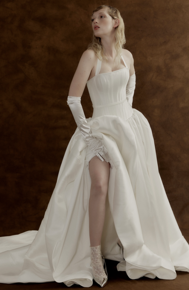 A woman in a white wedding dress with gloves lifting the skirt to reveal her leg, standing against a brown backdrop.