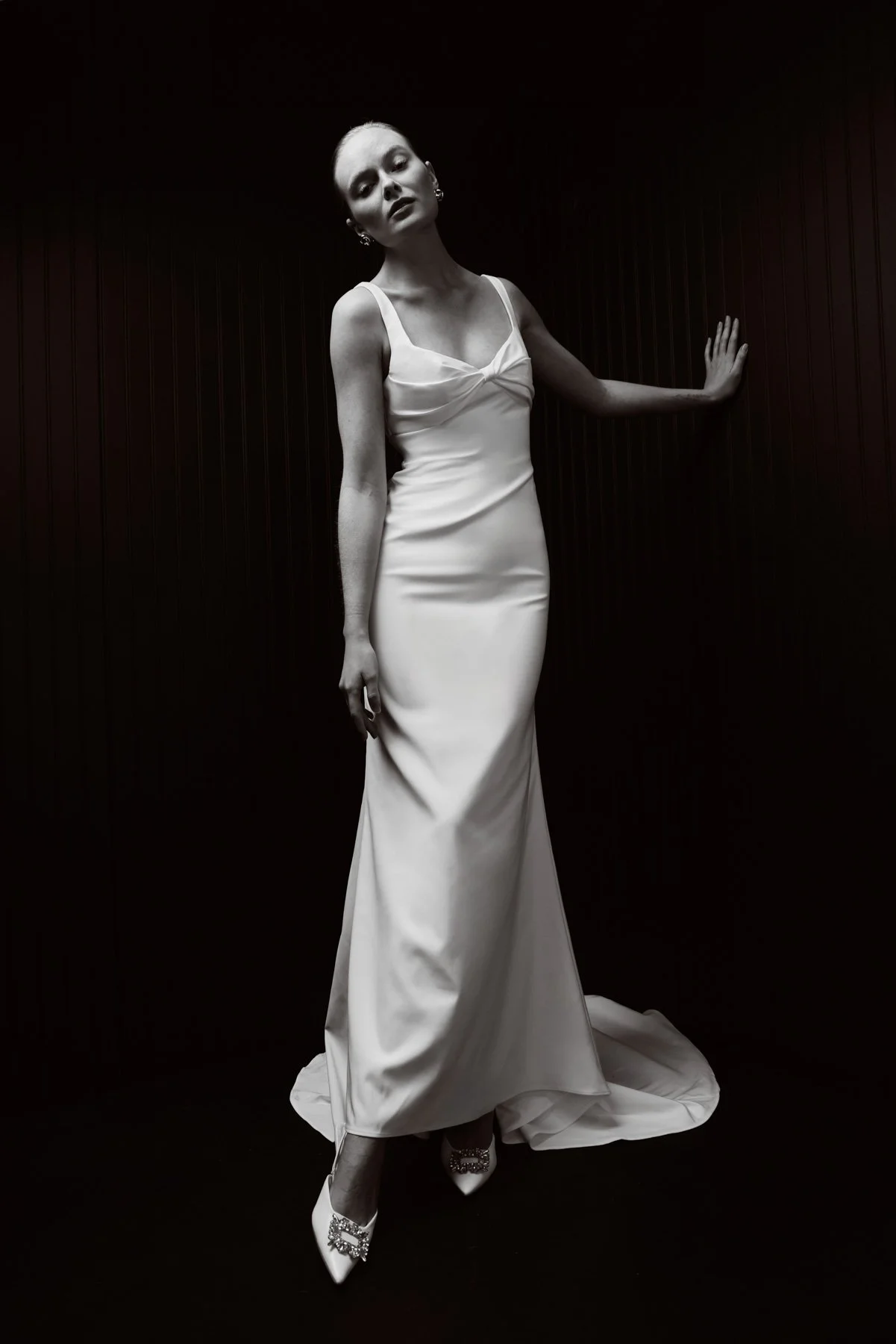 A woman in a white satin gown with spaghetti straps, standing against a dark background with her hand on the wall, looking at the camera.