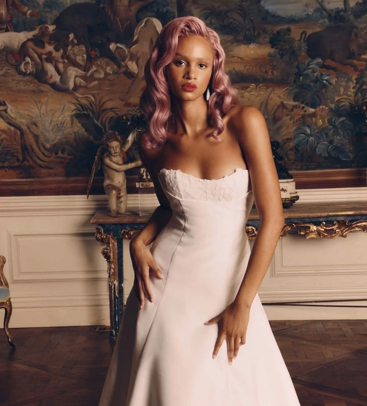 A woman with pink wavy hair standing in a room with ornate furniture and a detailed mural painting in the background.