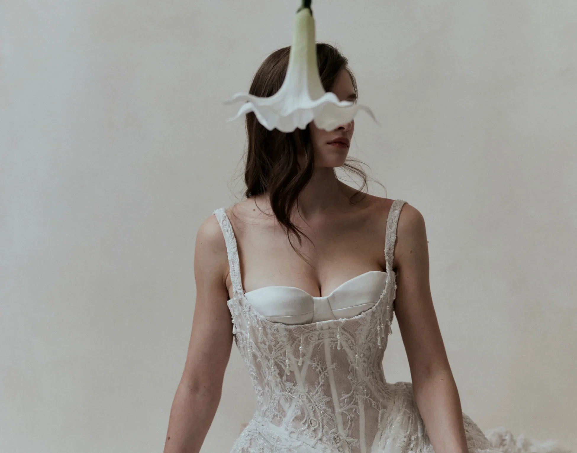 A woman in an elegant wedding dress with lace details and thin straps stands against a plain background. A white flower is suspended in front of her face, obscuring her eyes.