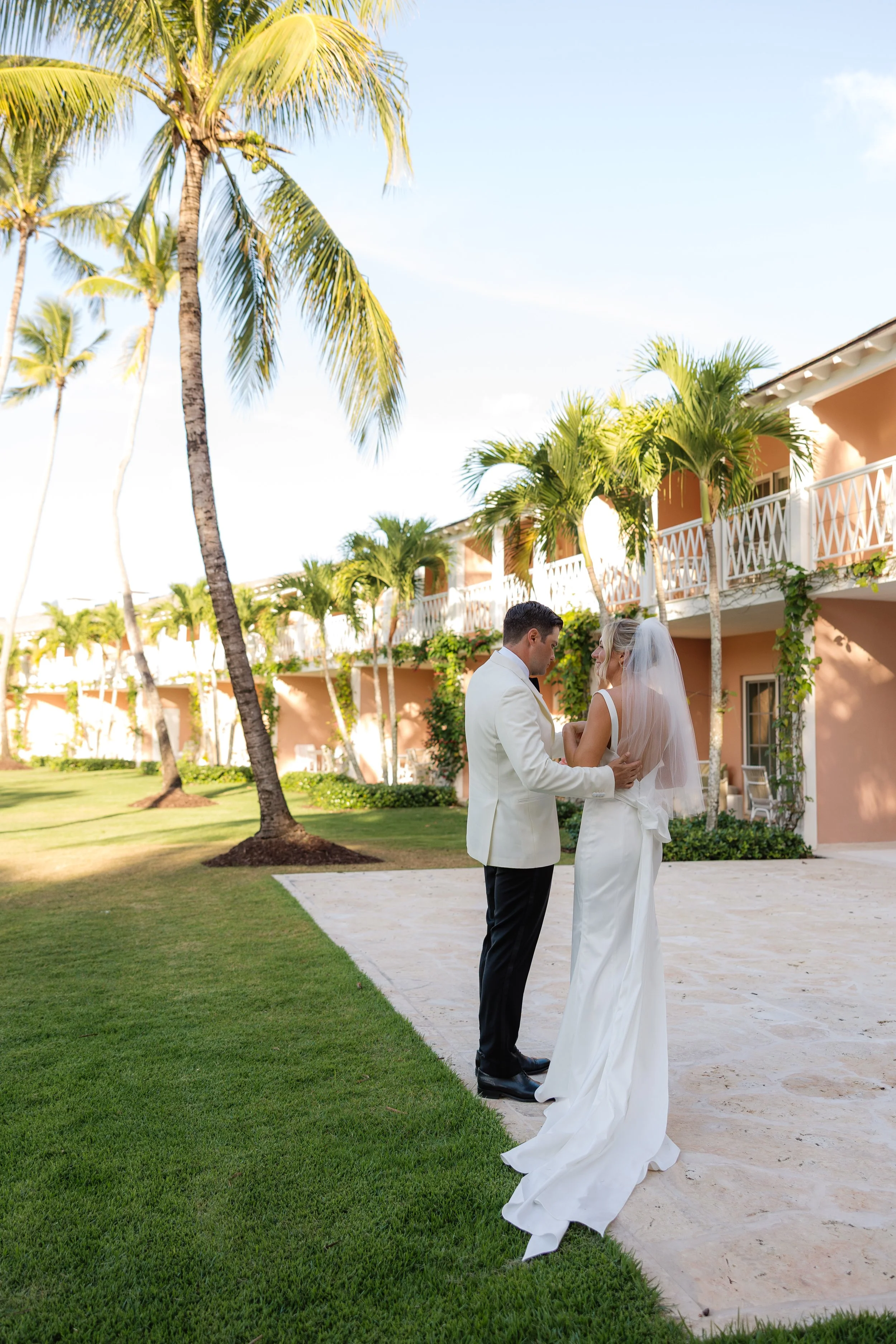 A bride and groom standing outdoors on a sandy walkway surrounded by green grass and tall palm trees, with a pink building featuring white balconies in the background, under a blue sky.