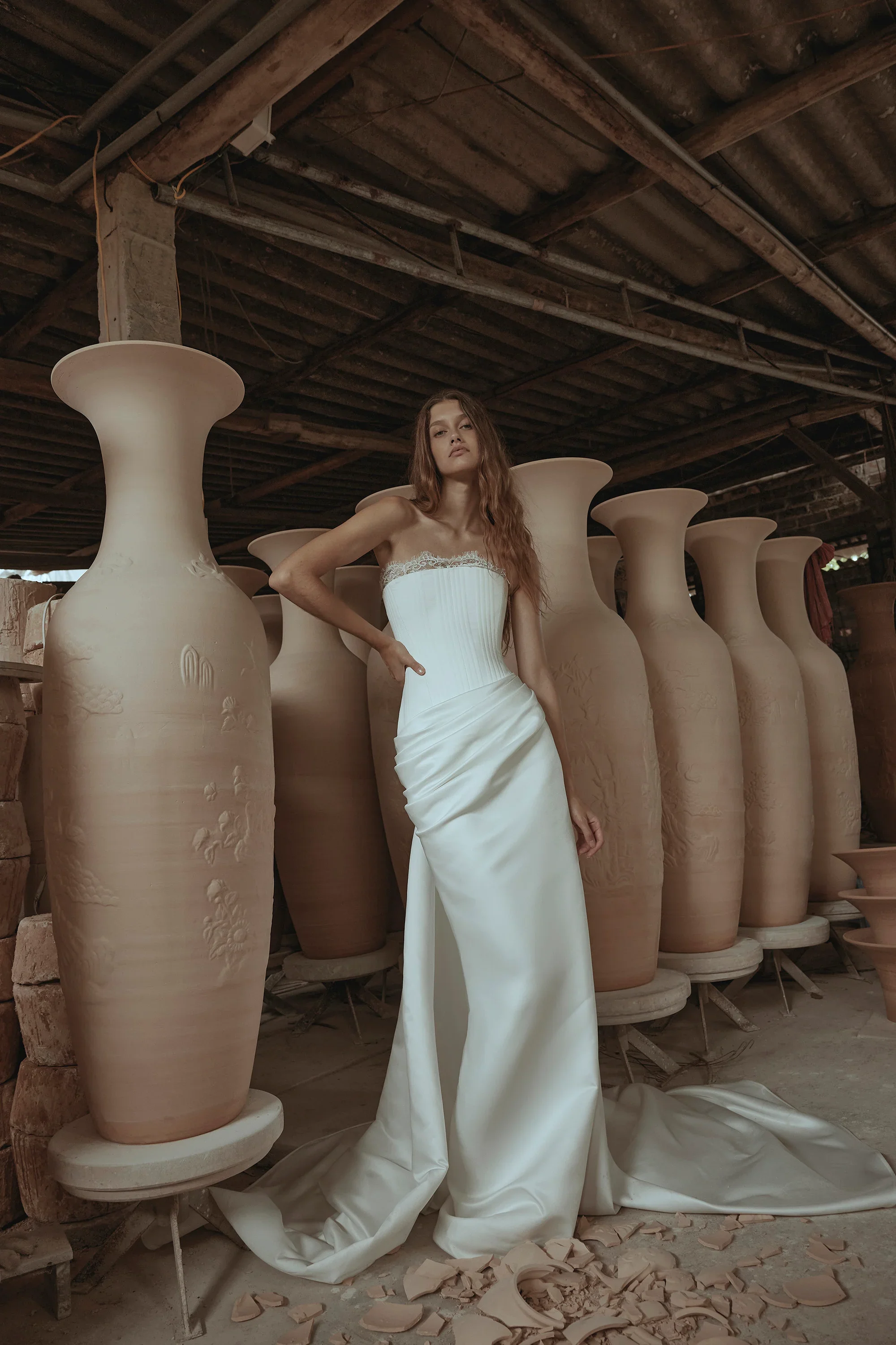 A woman in a white strapless wedding dress standing among large unglazed clay vases in a pottery studio.