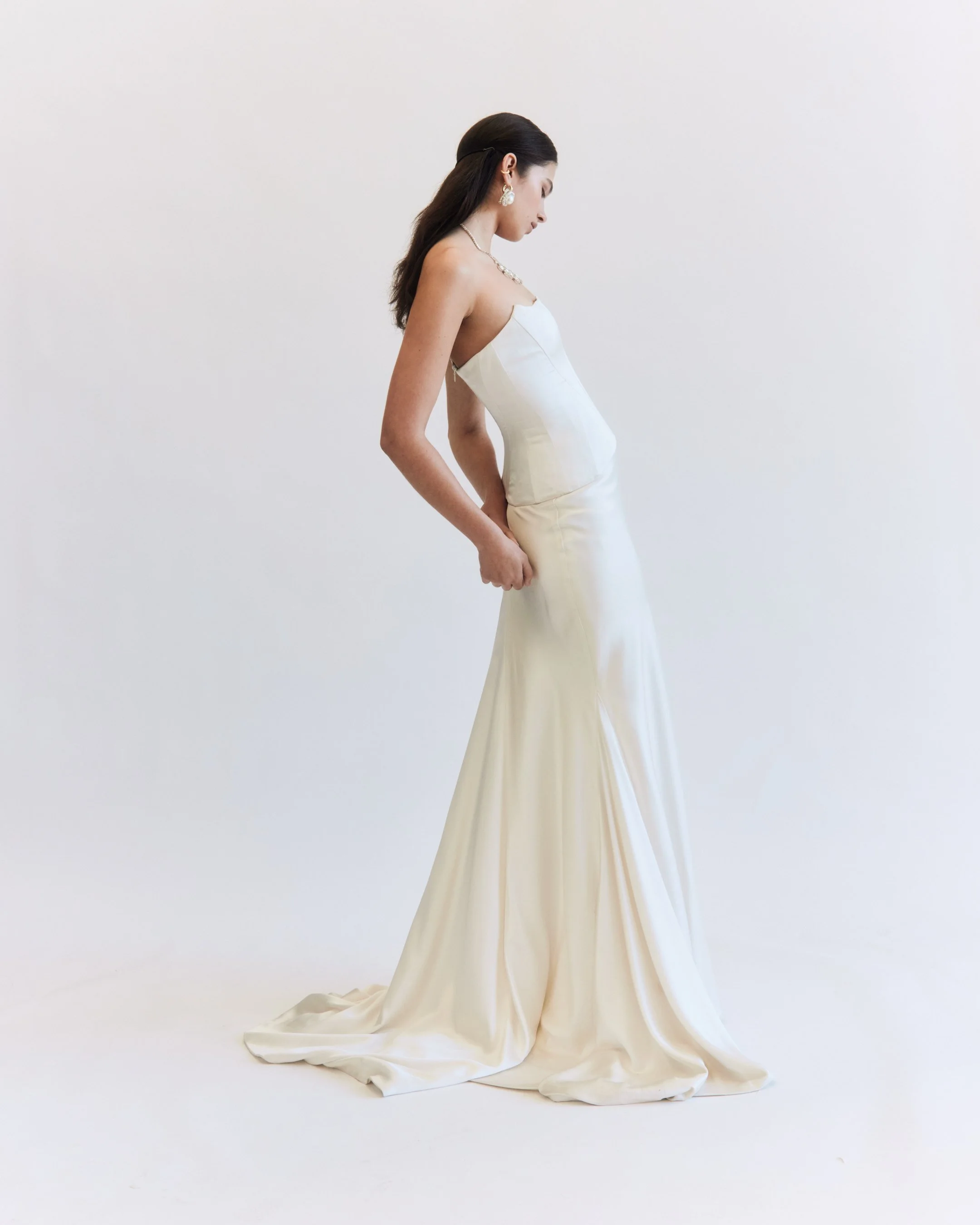 A woman in a white strapless gown stands against a plain white background, looking downward.