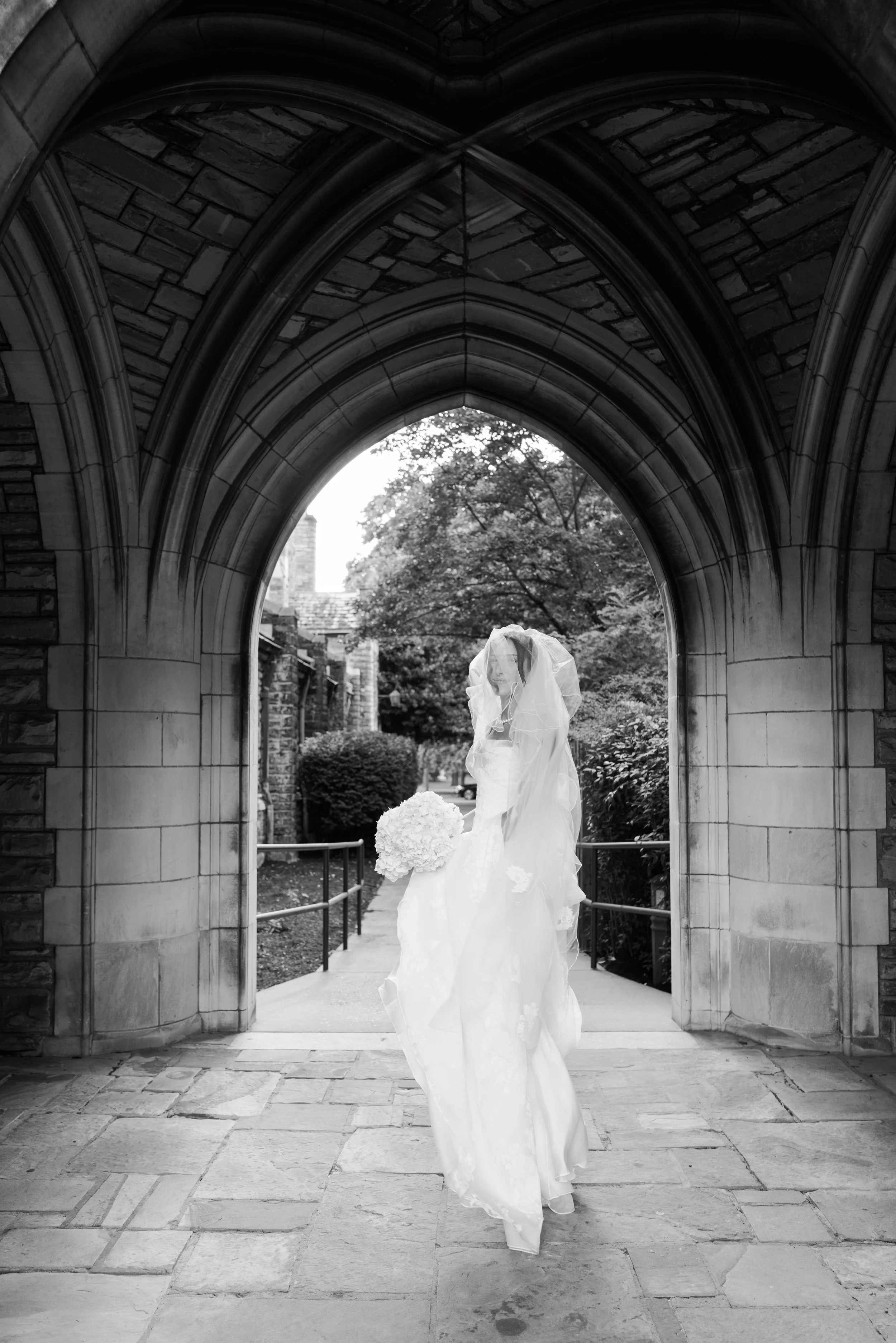 Black and white photo of a bride standing under a stone archway, holding a bouquet of flowers, with trees and buildings in the background.
