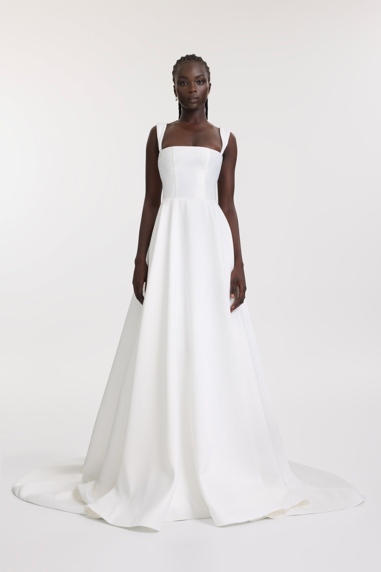 A woman wearing a long white formal gown with wide shoulder straps standing against a plain light background.
