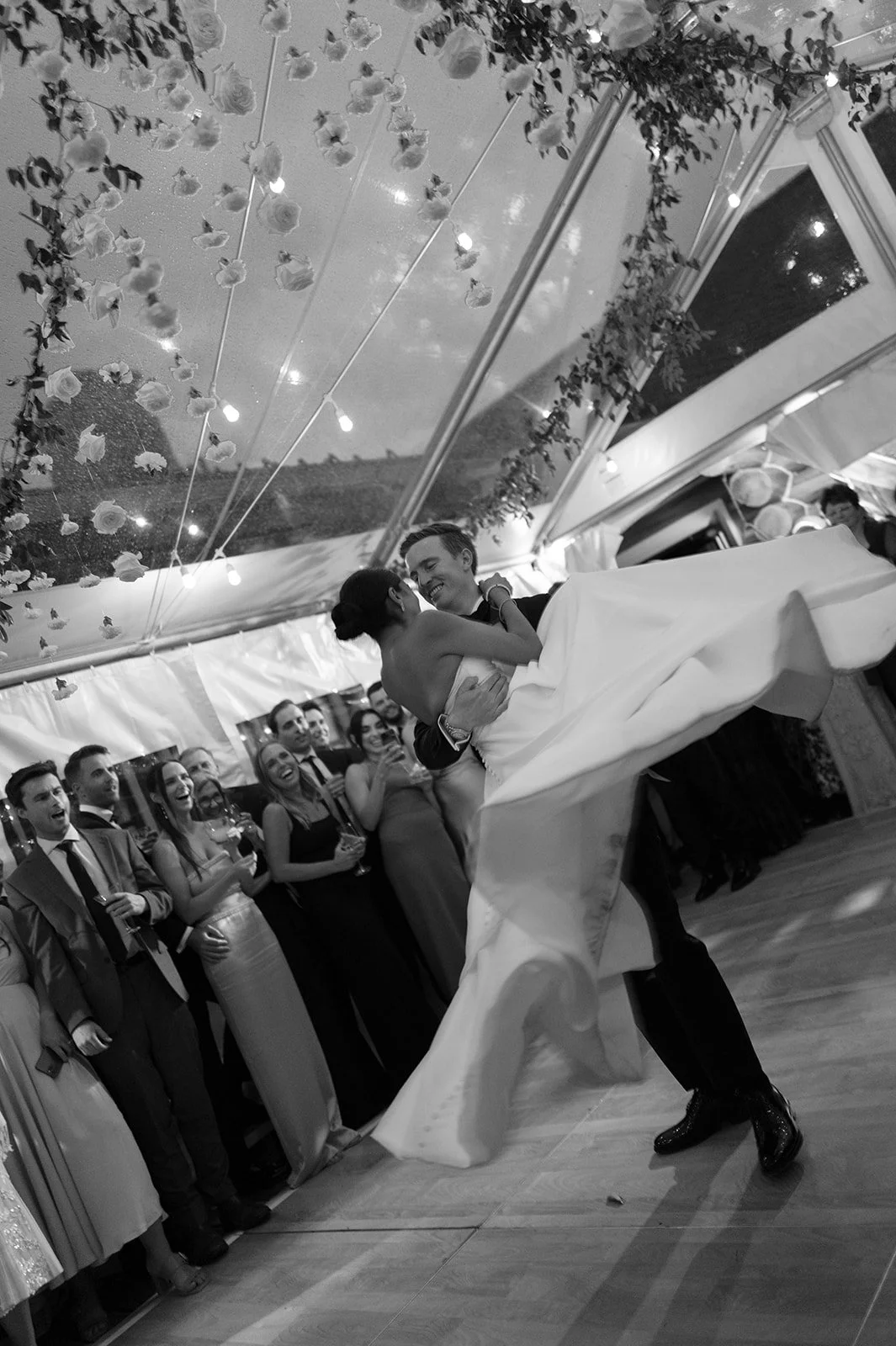 A couple dancing at a wedding reception, surrounded by guests, under a decorated tent with string lights and floral arrangements.