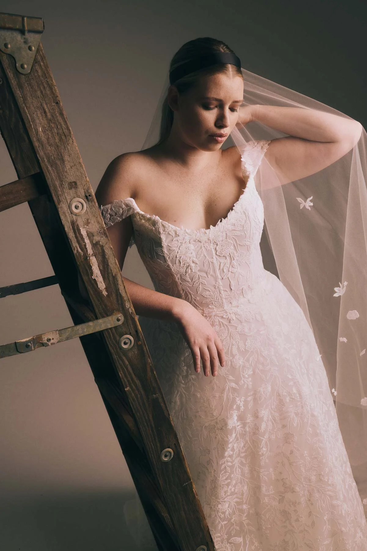 A woman in a lace wedding dress with a veil stands next to a wooden ladder, touching her forehead with her eyes closed.