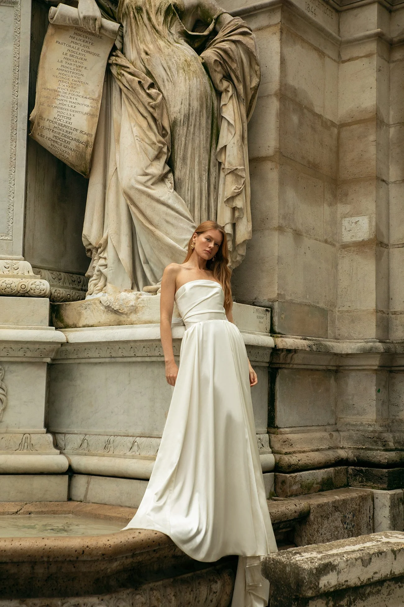 A woman in a strapless white gown standing next to a fountain and a large stone statue on a historical building.