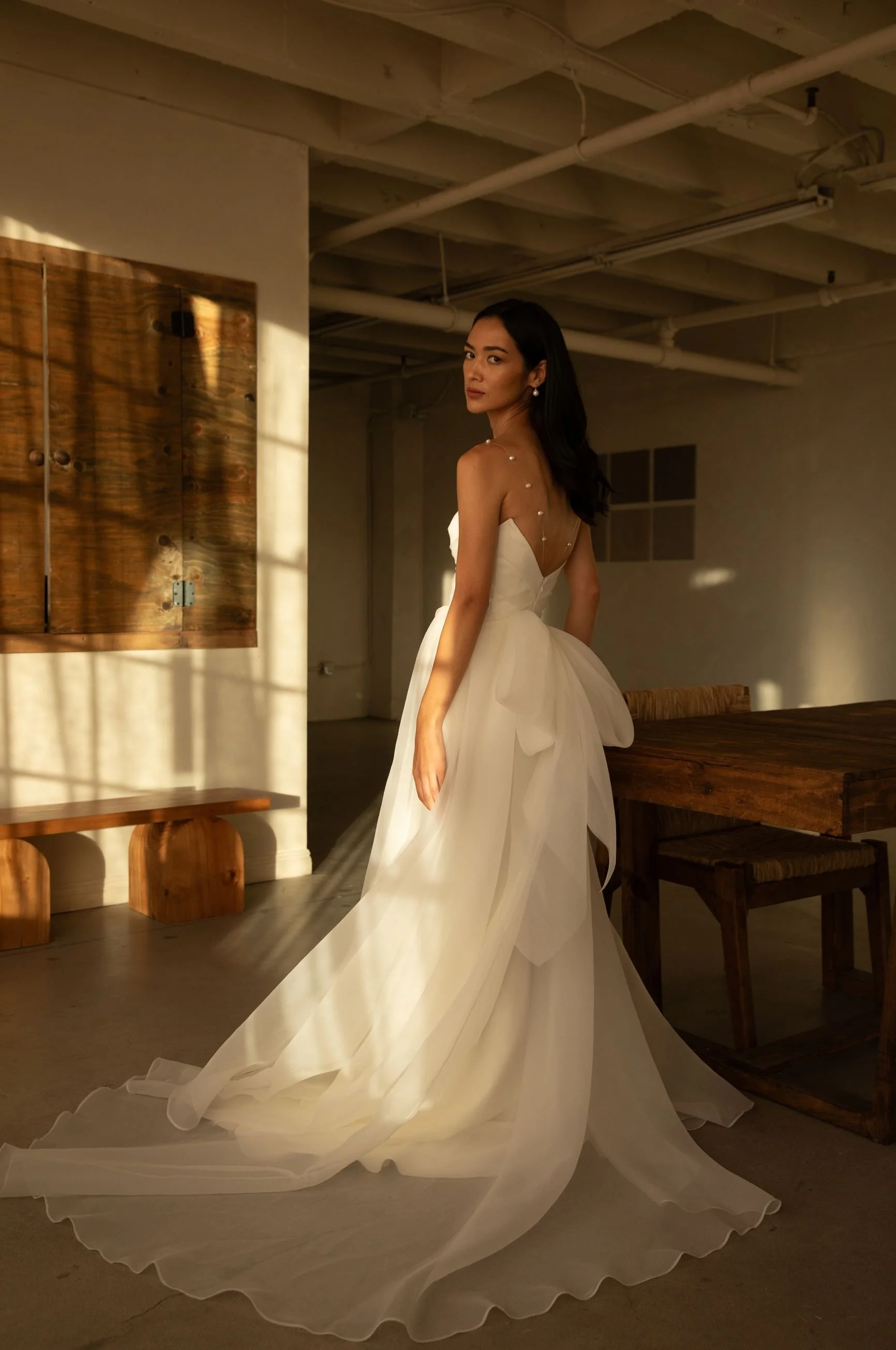 A woman in a white wedding dress with a train, standing in a room with warm sunlight, wooden furniture, and an industrial-style ceiling.