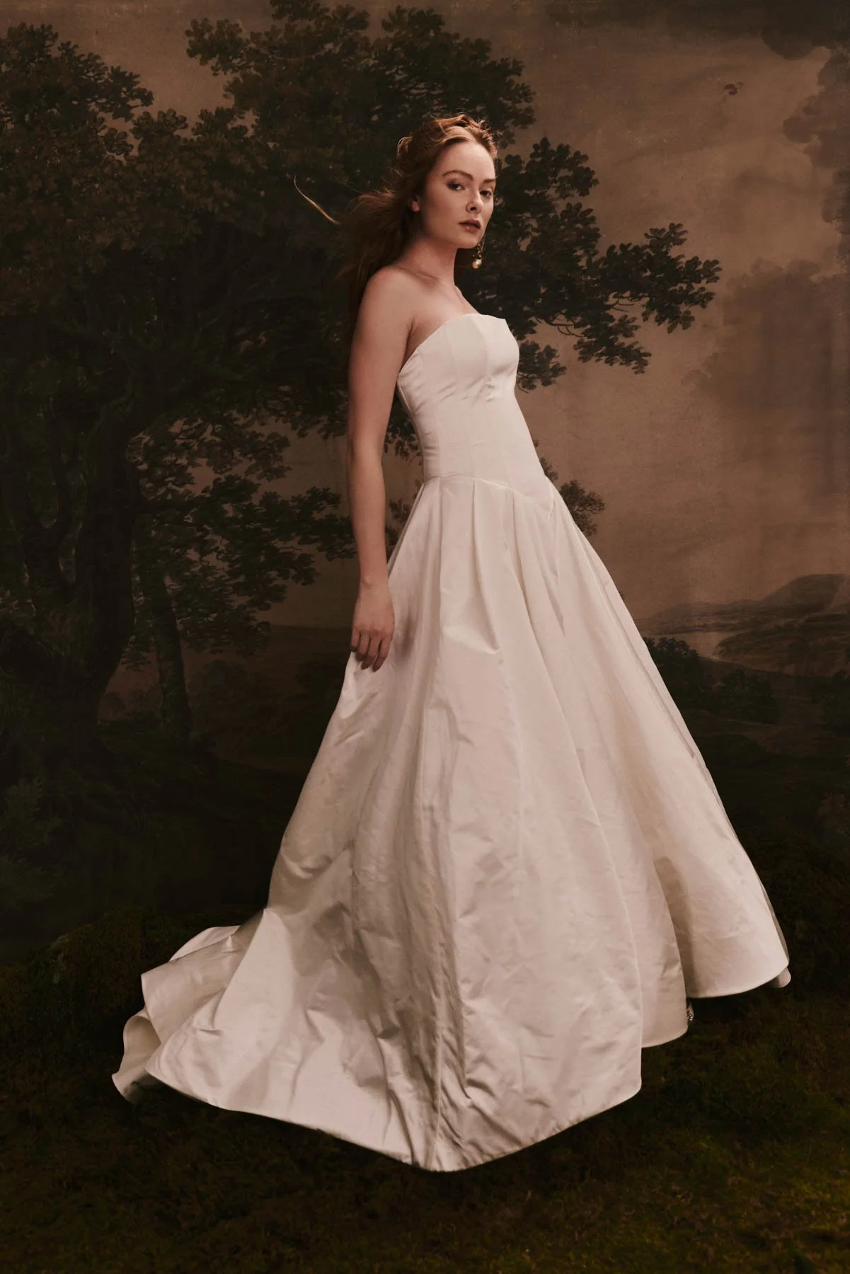 A woman in a strapless white gown standing outdoors against a painted backdrop of trees and sky.